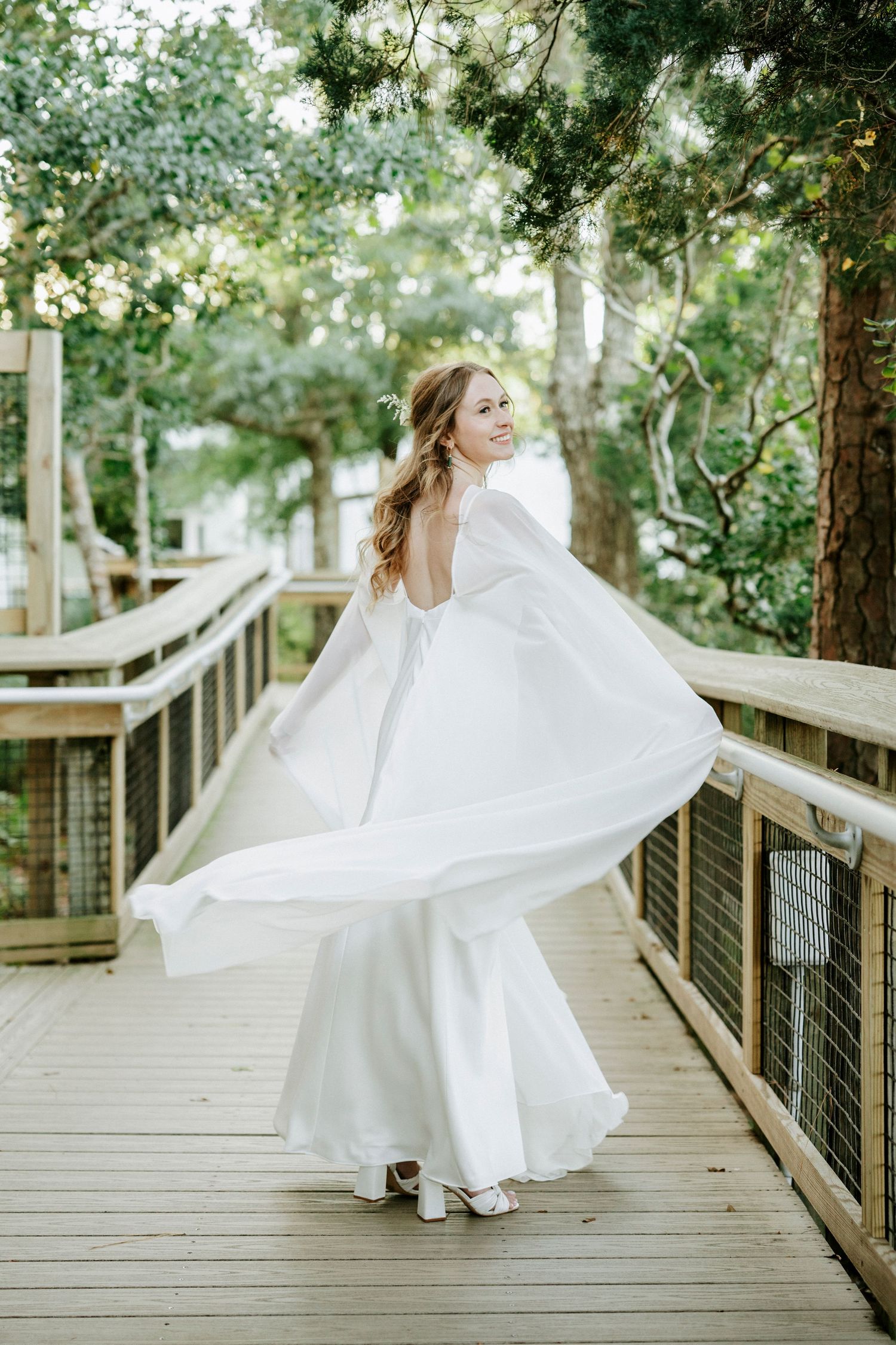A white wedding dress spins elegantly on a wooden deck pathway amidst natural greenery.