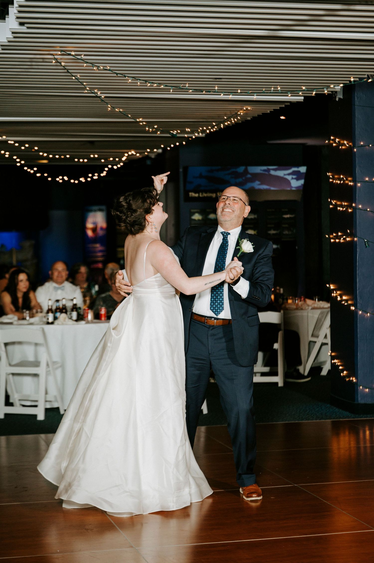 A couple shares a dance at a wedding reception under twinkling string lights in an indoor venue.