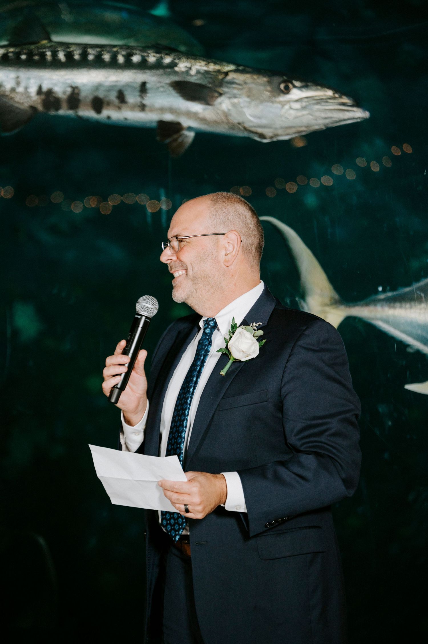 A speaker in a dark suit gives a speech at an evening wedding reception.