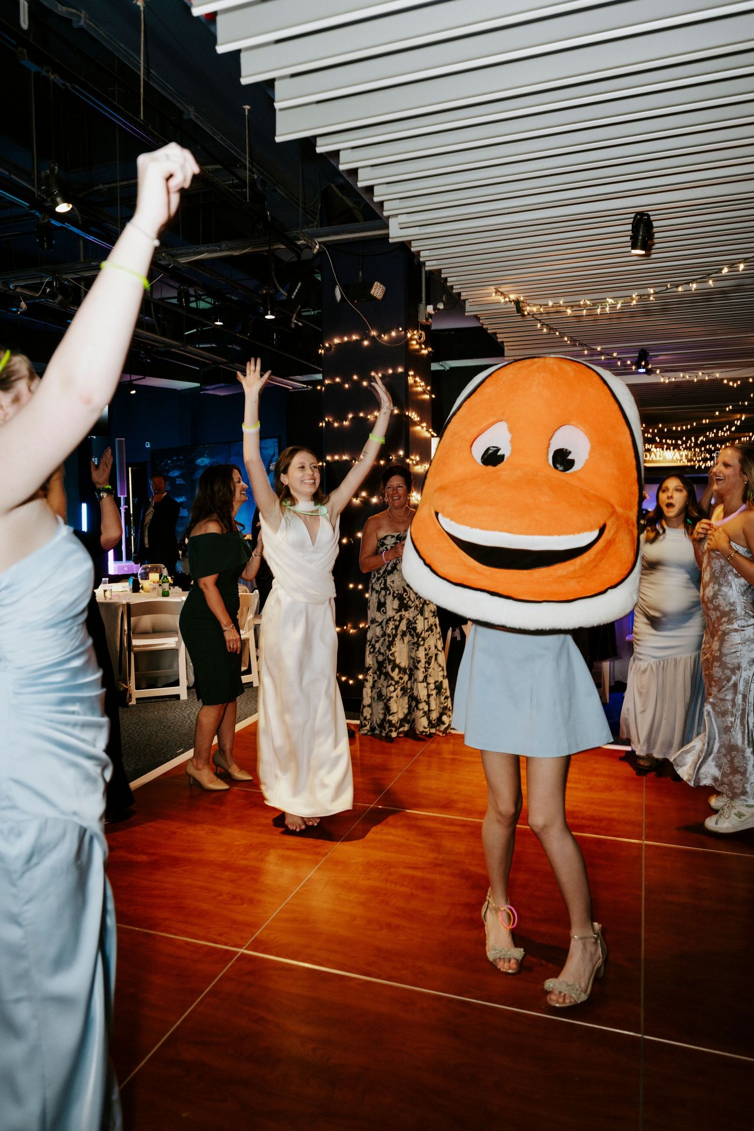Wedding guests dance and celebrate on a wooden dance floor under string lights at an indoor reception venue.