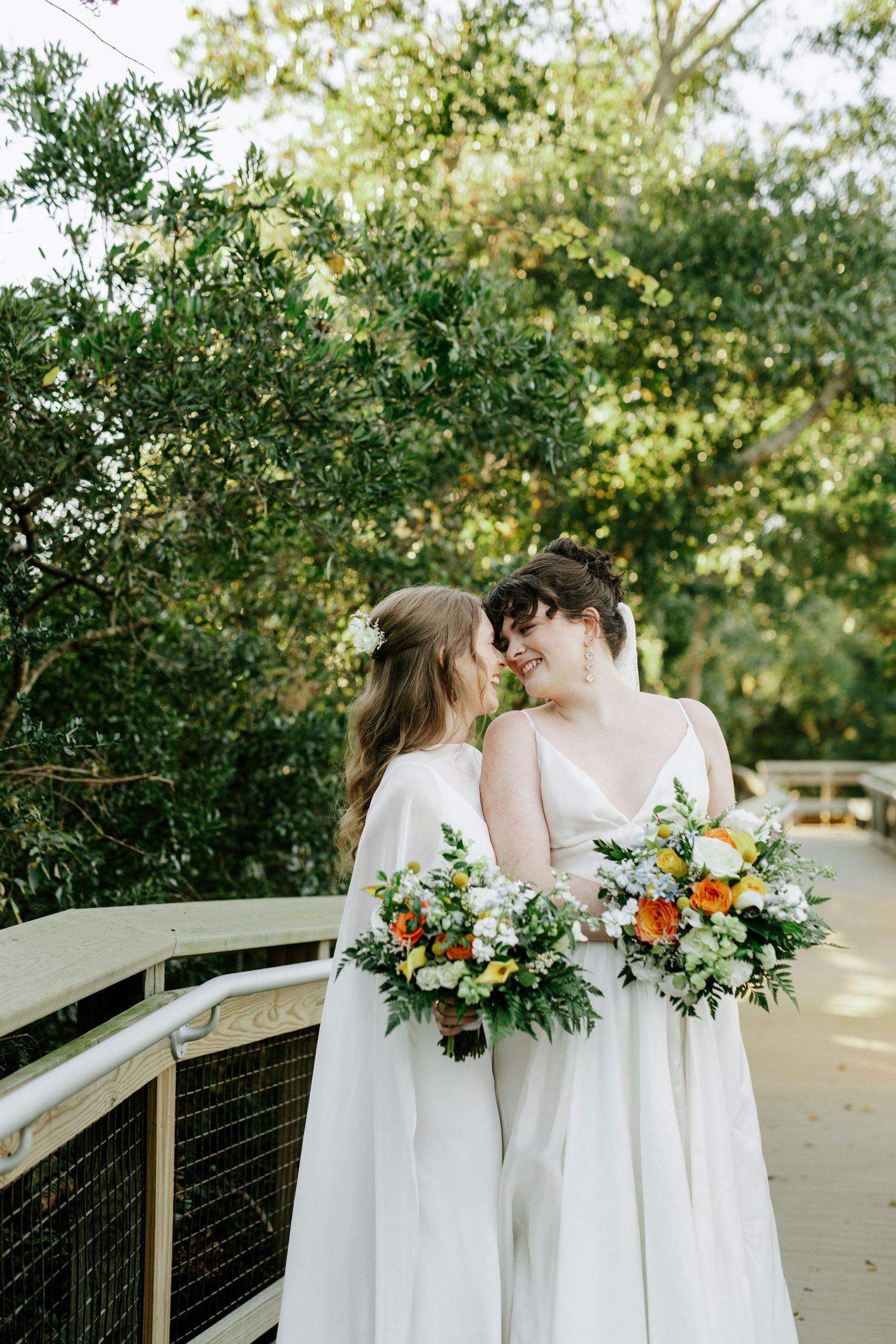 Two people in white wedding dresses share a tender moment on a wooden boardwalk surrounded by trees.