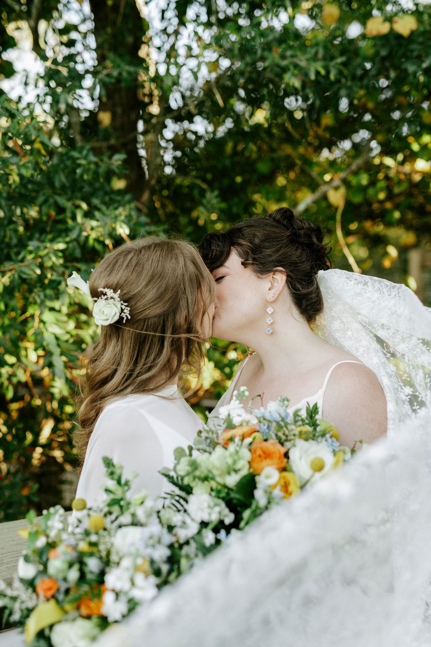 Two brides share a tender moment in a garden setting with vibrant orange and white wedding bouquets.
