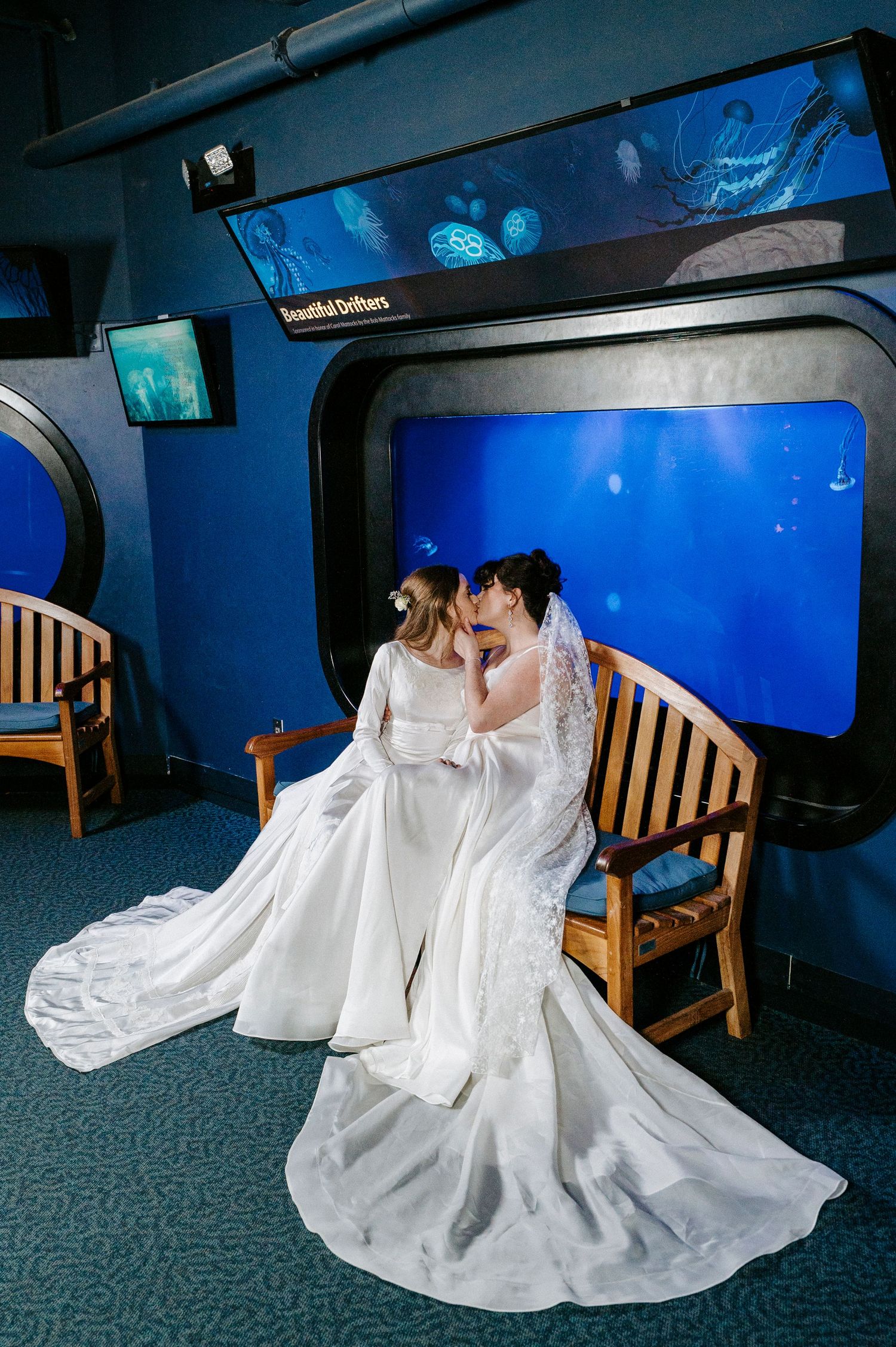 Two people in white wedding dresses sit together on a wooden bench against a deep blue aquarium backdrop.