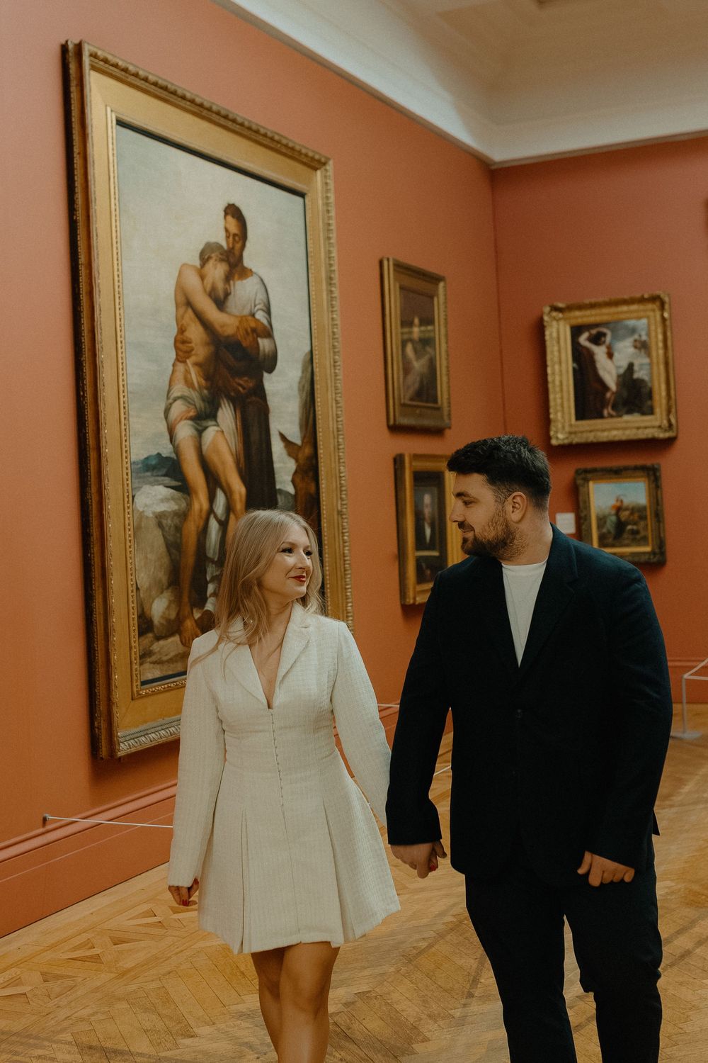 A couple walks through an art gallery with orange walls displaying framed paintings in multiple connected shots.