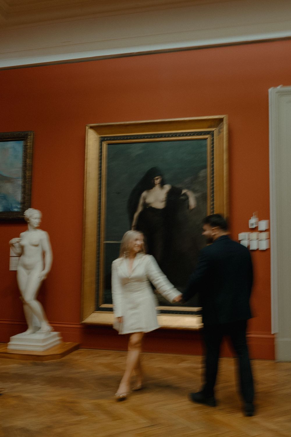 Two people walk through an art gallery with orange walls displaying classical paintings and a white marble statue.
