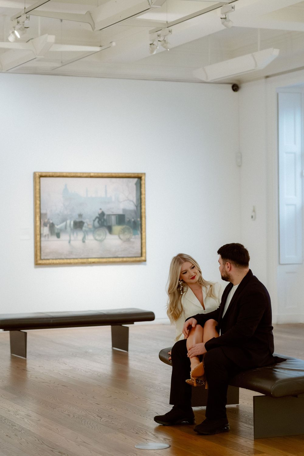 Two people sit together on a bench in a minimalist art gallery with framed landscape paintings on white walls.