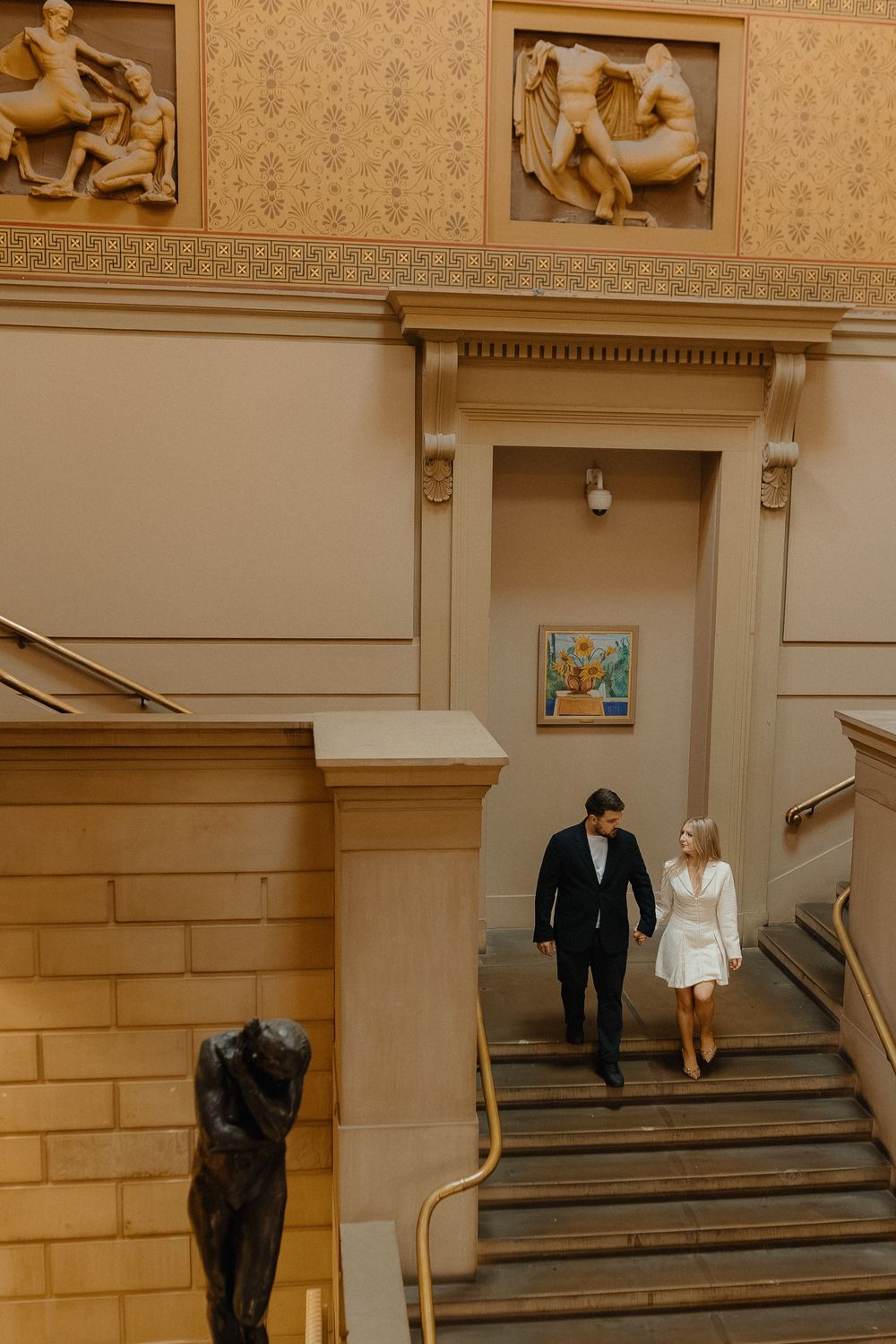 Two people in formal attire descend a grand marble staircase in an elegant indoor setting.