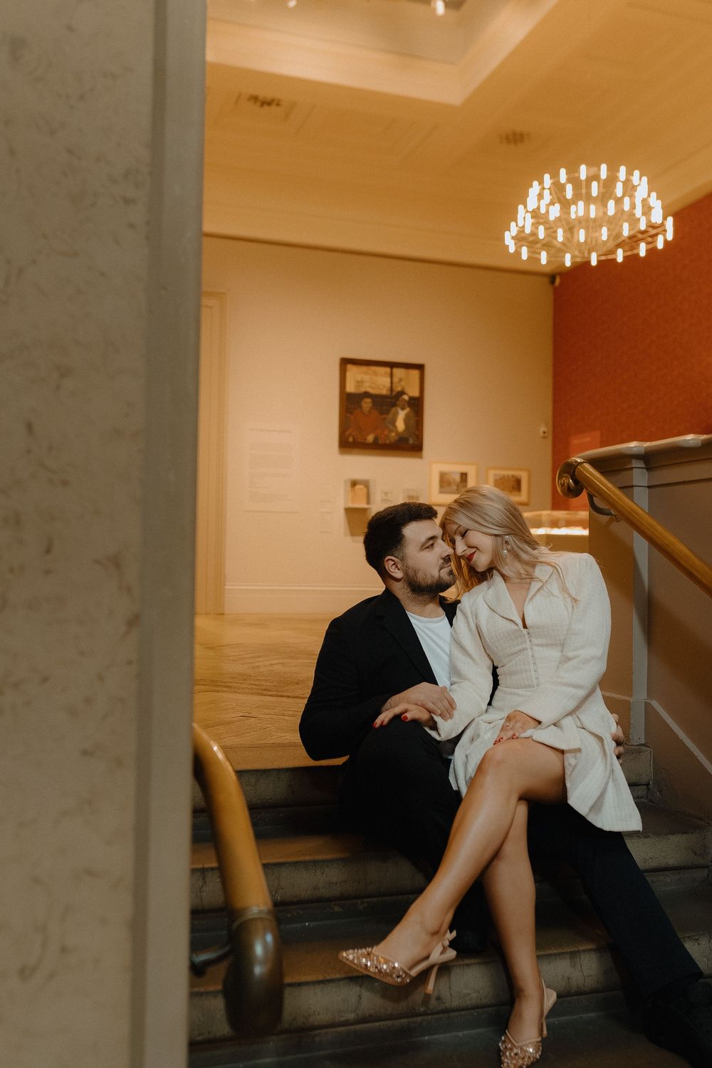 A romantic couple sits together on elegant marble stairs in an upscale venue with warm lighting and chandeliers.