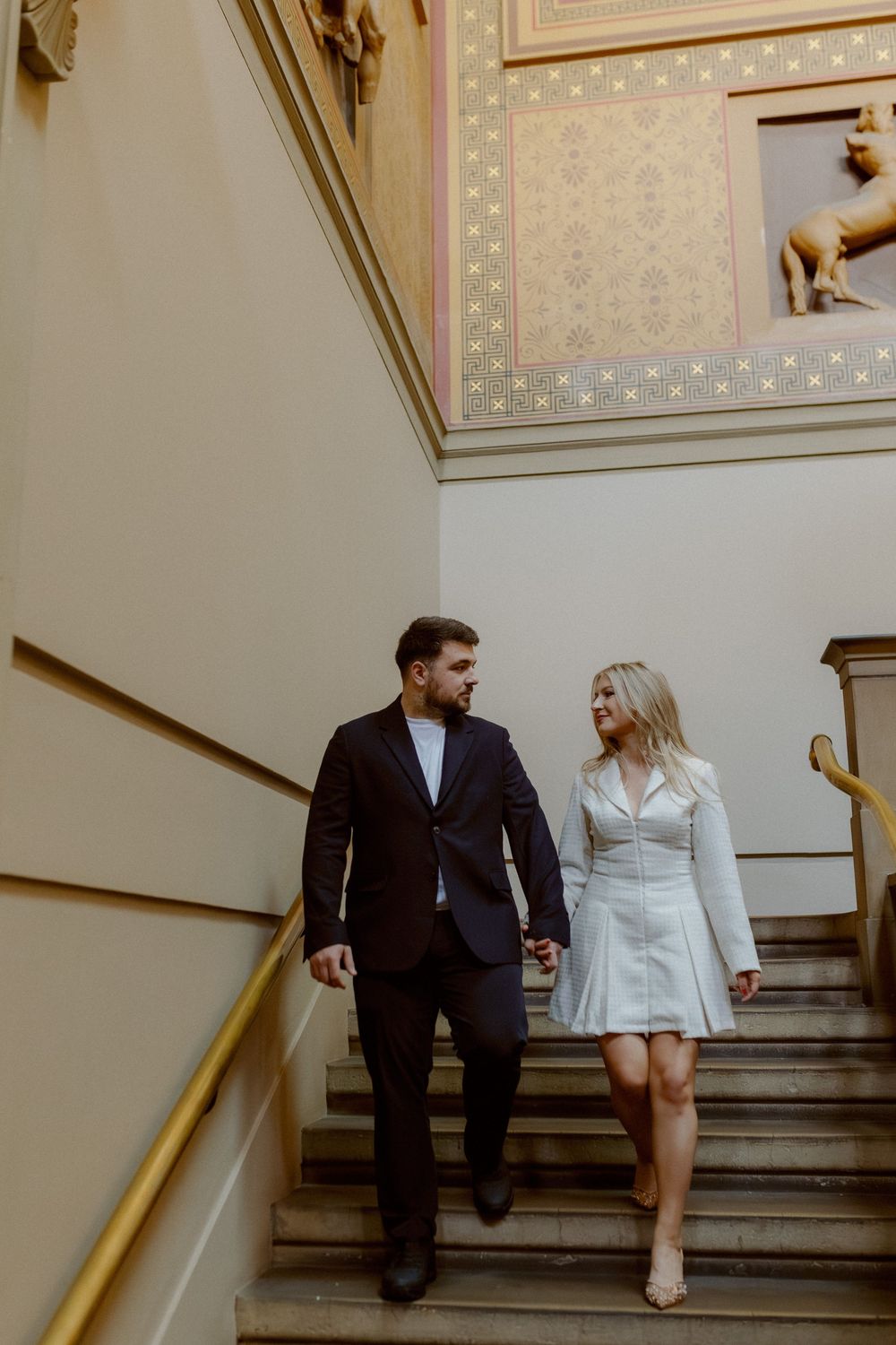 A couple shares an intimate moment while seated on grand marble stairs in a classic interior setting.