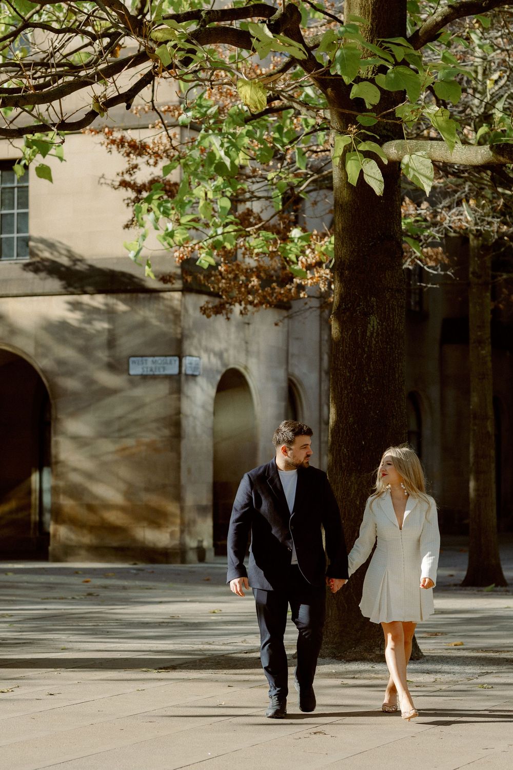 A couple in formal attire walks together under green trees in a romantic outdoor setting.