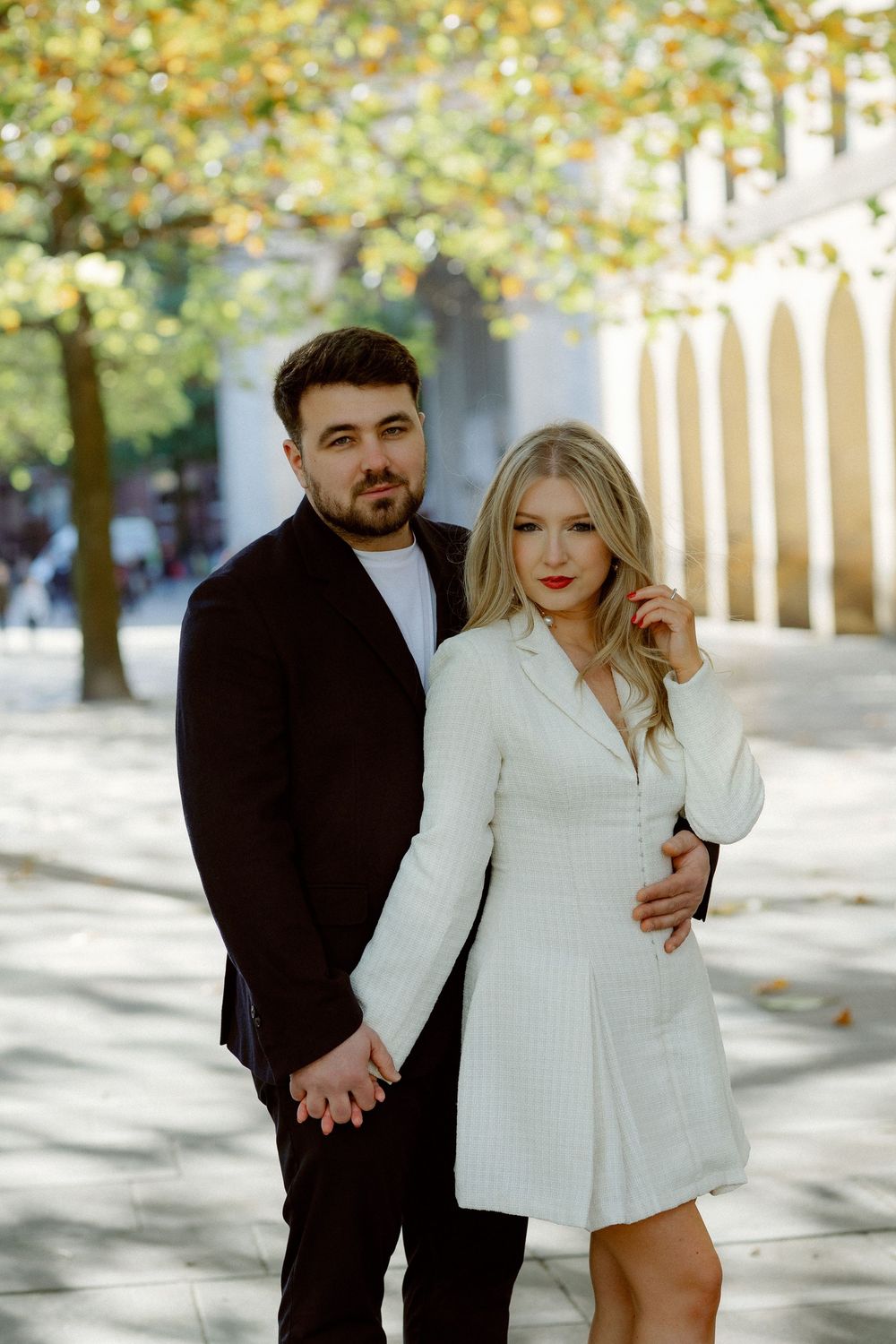 A romantic couple in white and black outfits poses together in a sunlit outdoor setting with autumn trees.