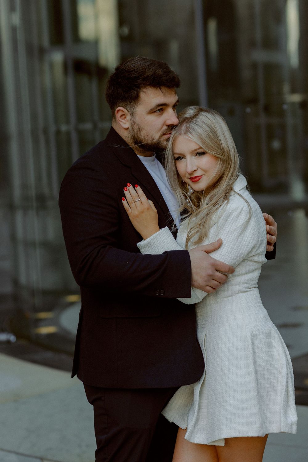 A couple shares an intimate moment against an urban backdrop with modern architecture and natural lighting.