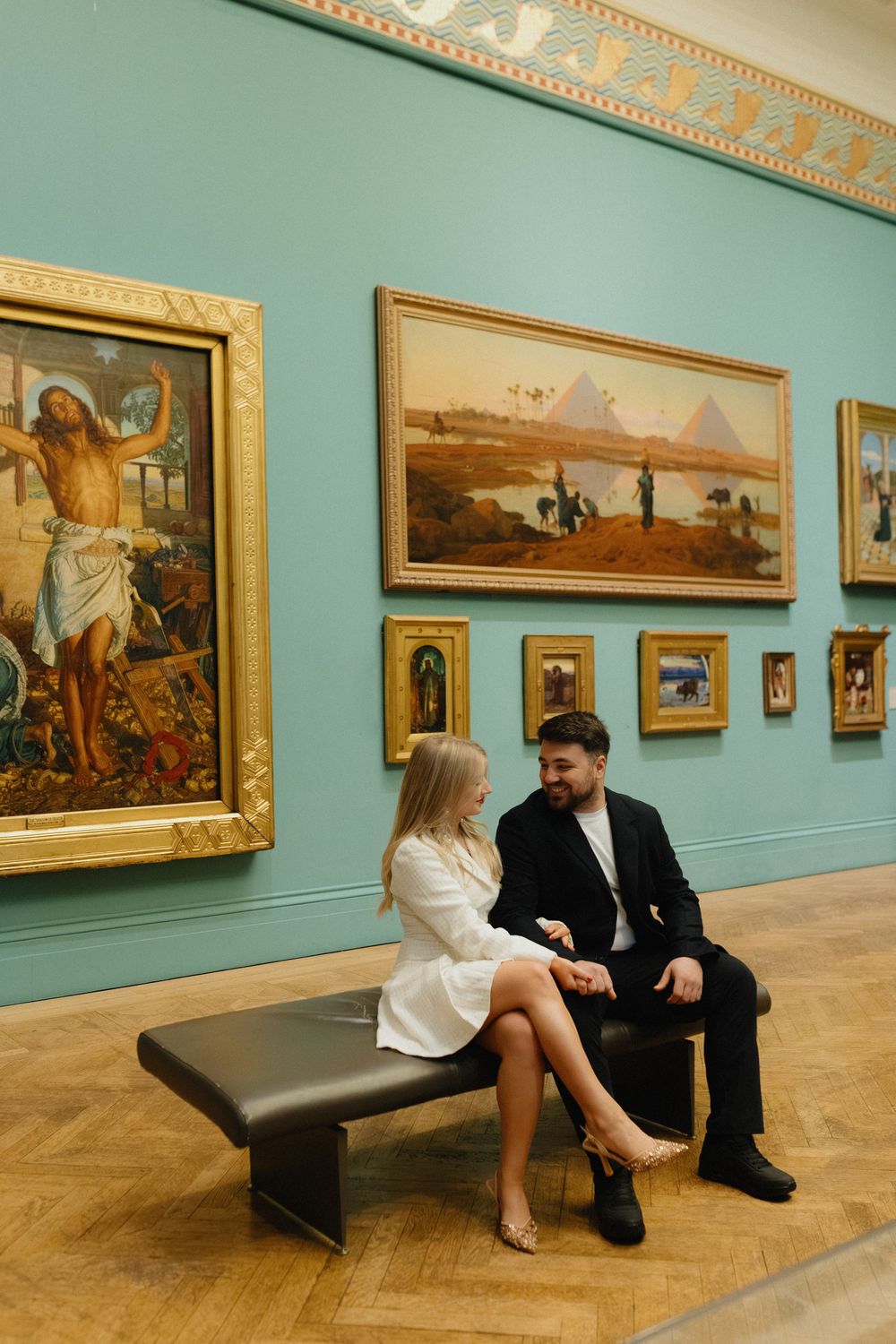 Two people sit on a modern bench in an art museum gallery with ornate gold-framed paintings on turquoise walls.