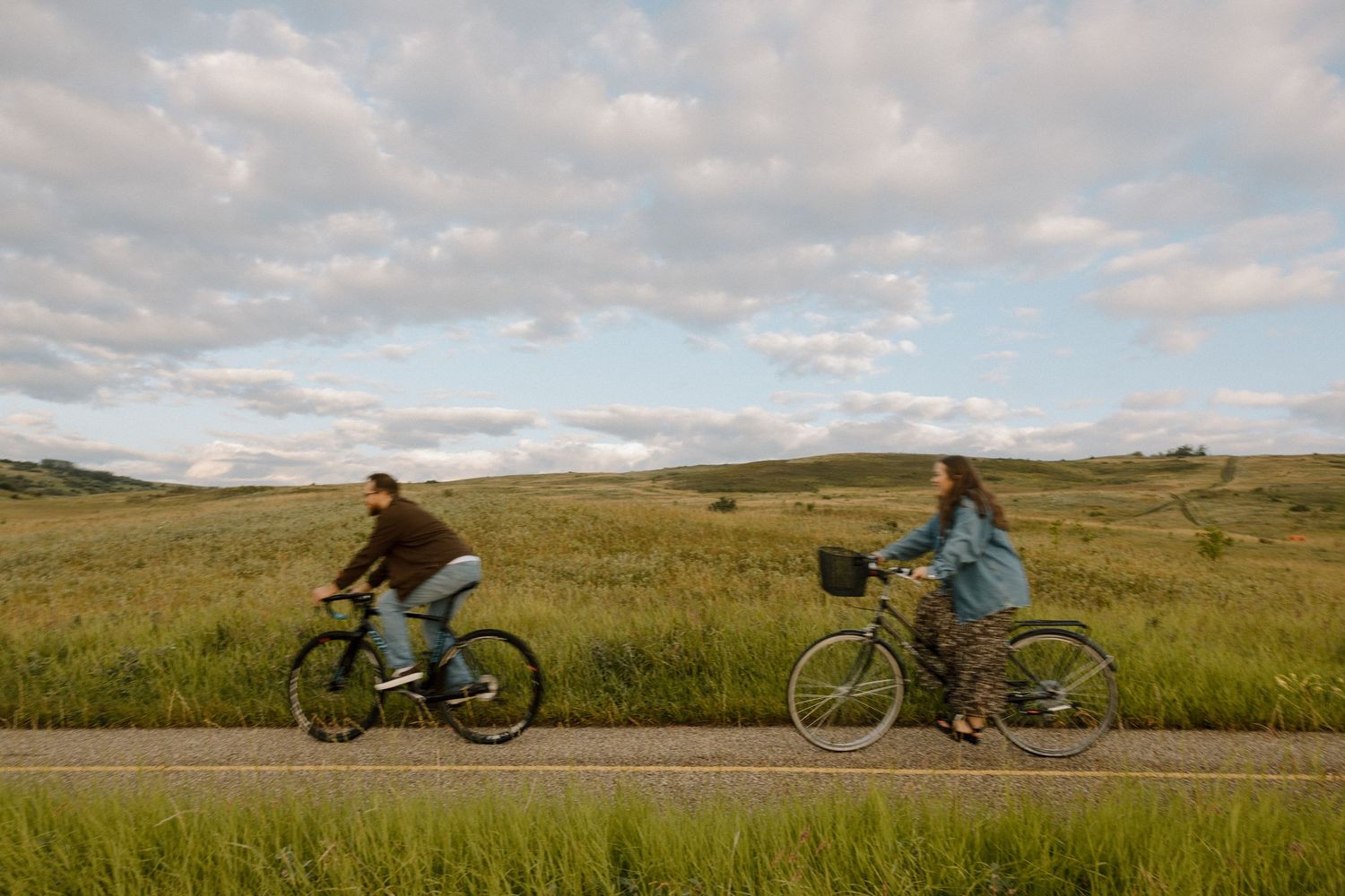 Cyclists ride their bikes along a scenic mountain road through vast rolling grasslands under a cloudy sky.