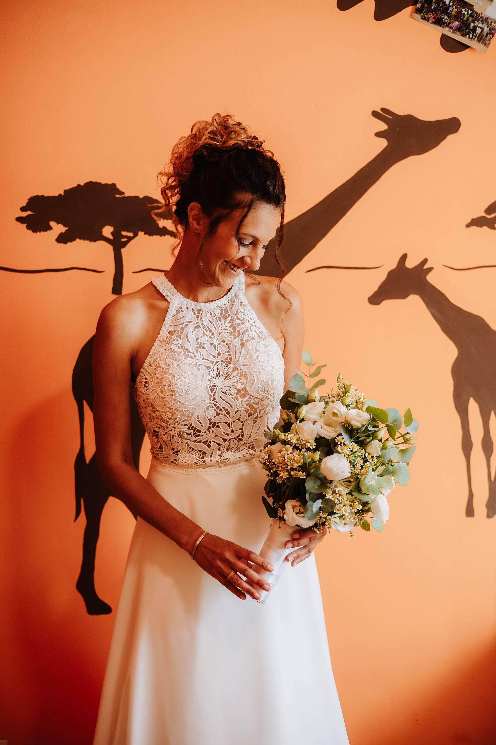 A bride in a white lace wedding dress holds a bouquet against an orange wall with shadow silhouettes.