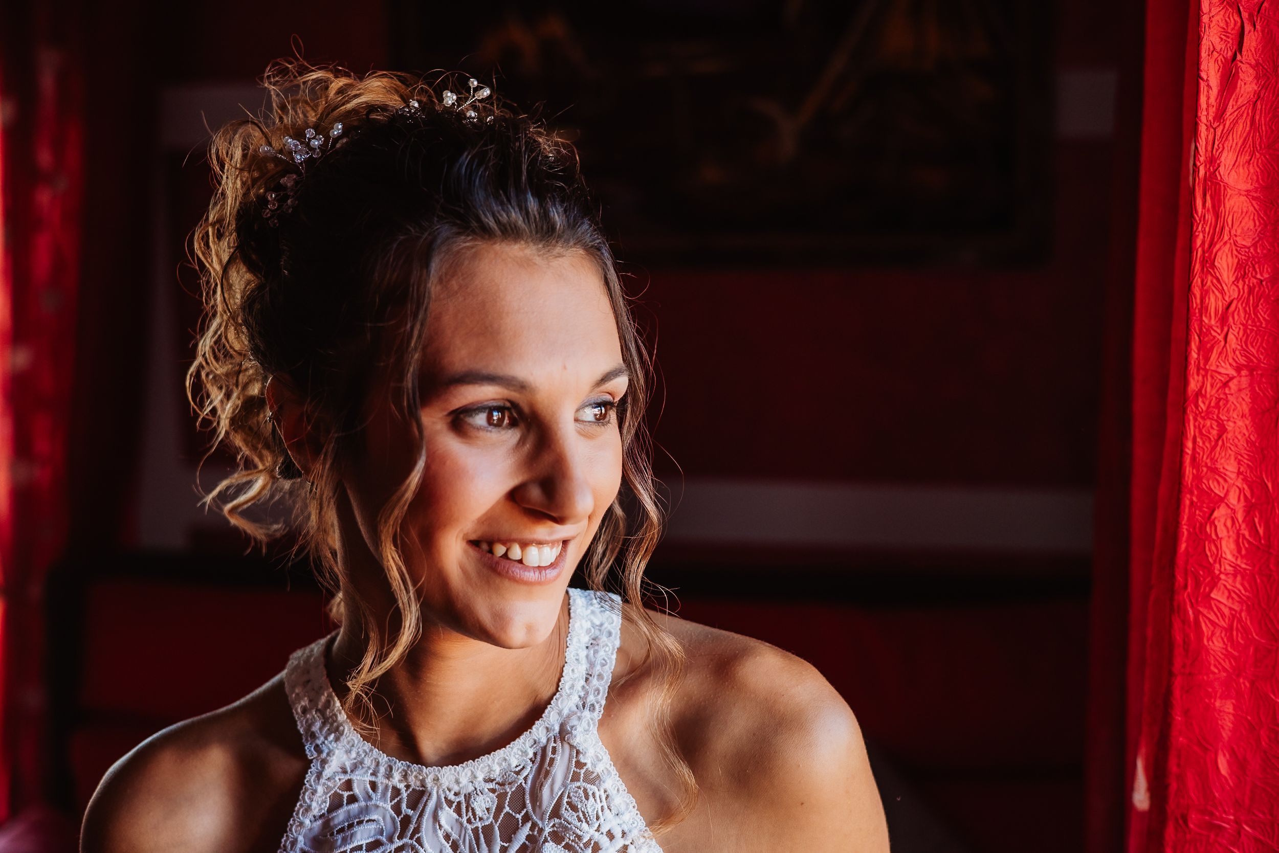 A smiling woman in a white lace halter top poses against a dark background with red curtain.