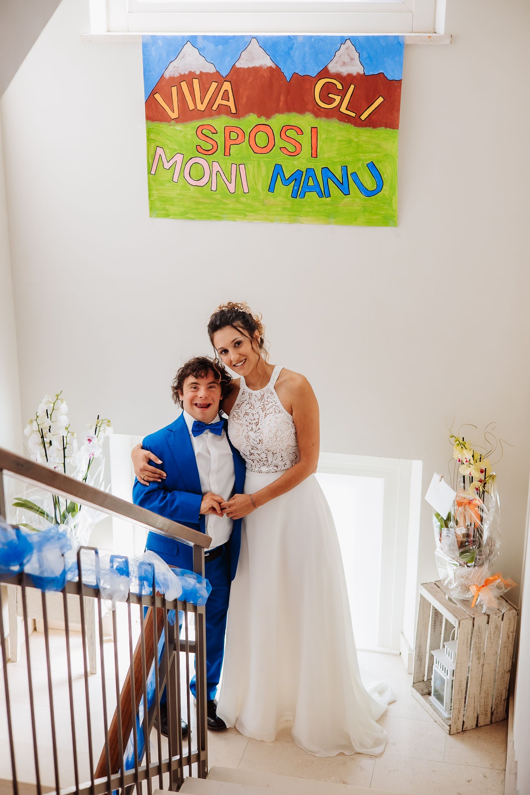 A couple stands on a staircase under a colorful banner reading 'Viva gli sposi mani mano' at a wedding celebration.