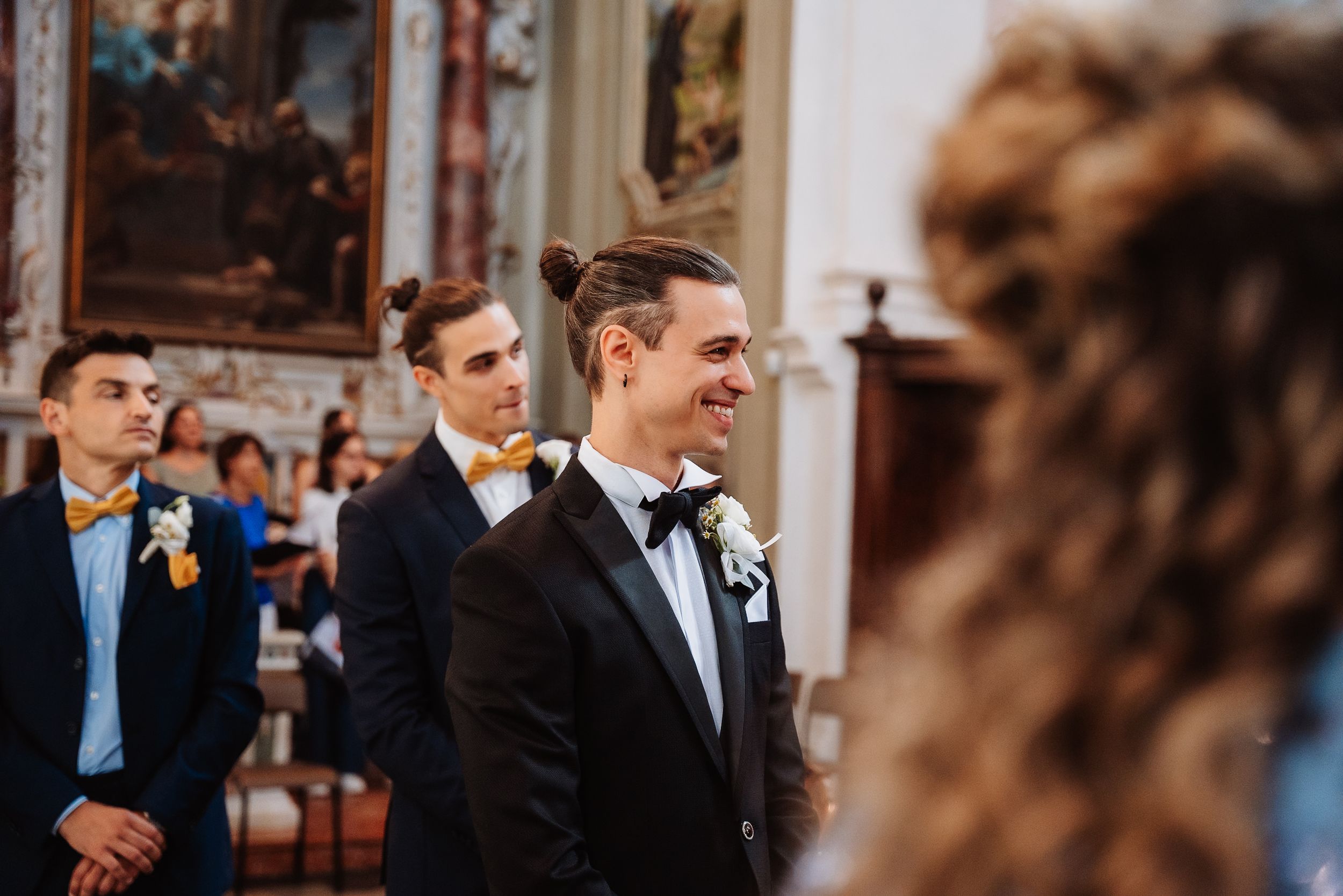 A groom in a black tuxedo and bow tie smiles joyfully during a wedding ceremony in an ornate church setting.