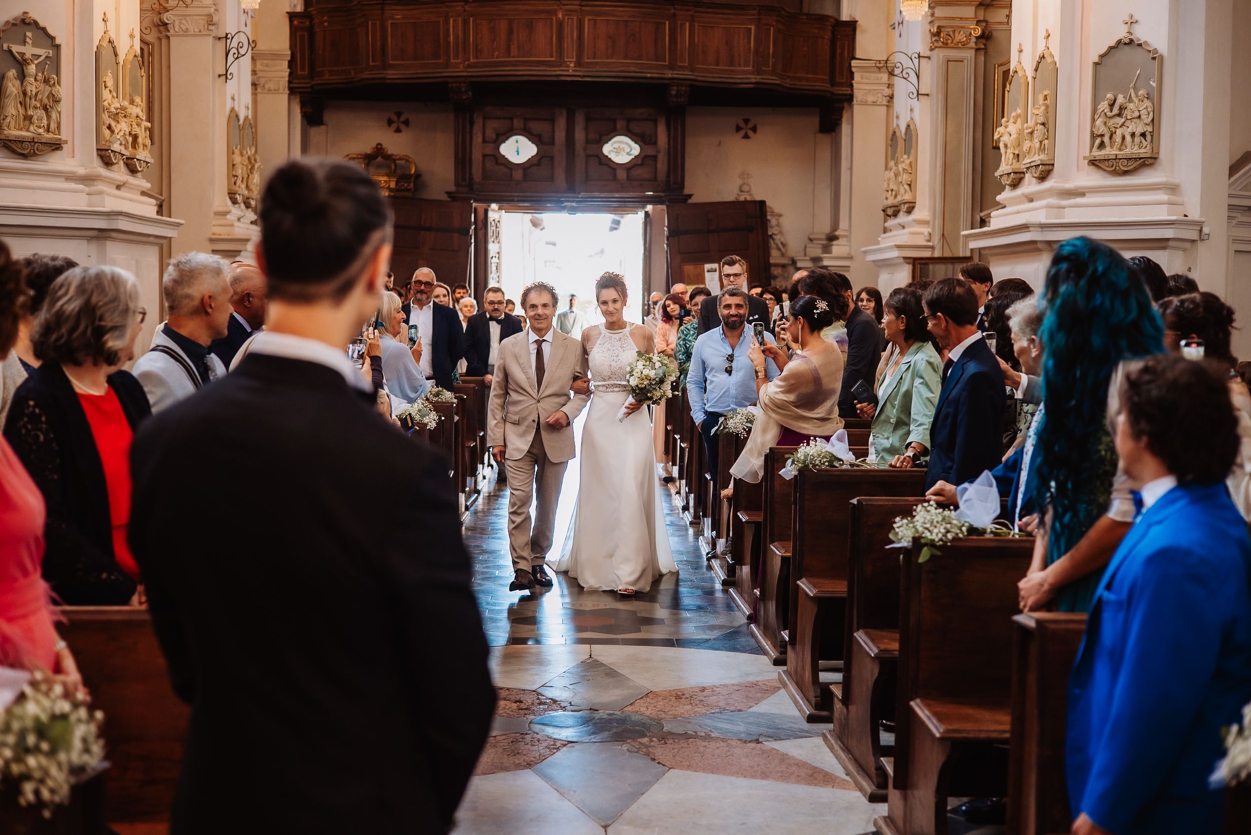 Wedding ceremony guests stand in church pews as bride and groom walk down ornate aisle with arched ceilings.