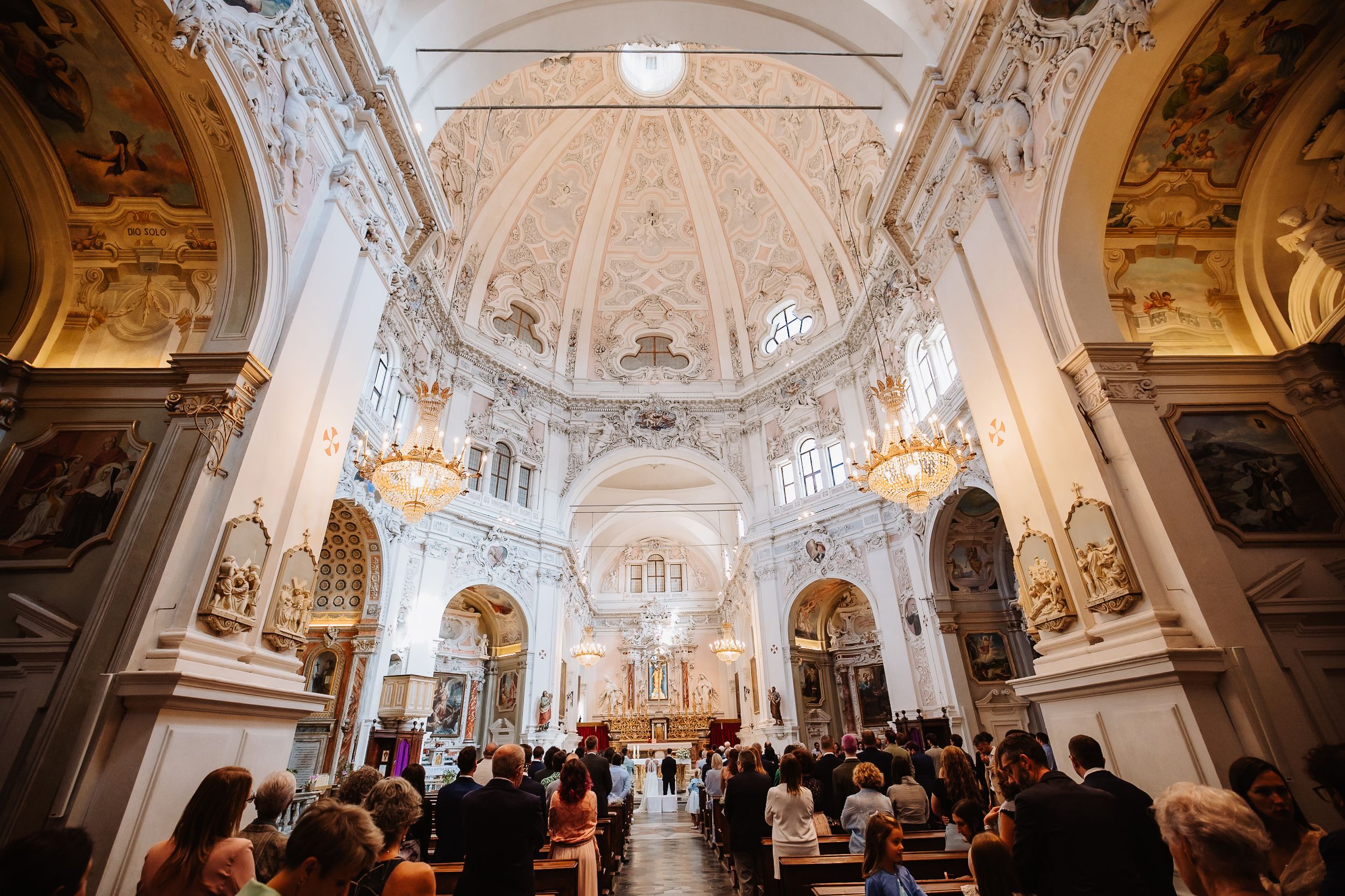 Panoramic view of an ornate baroque church interior with white and gold decor, vaulted ceilings, and chandeliers.