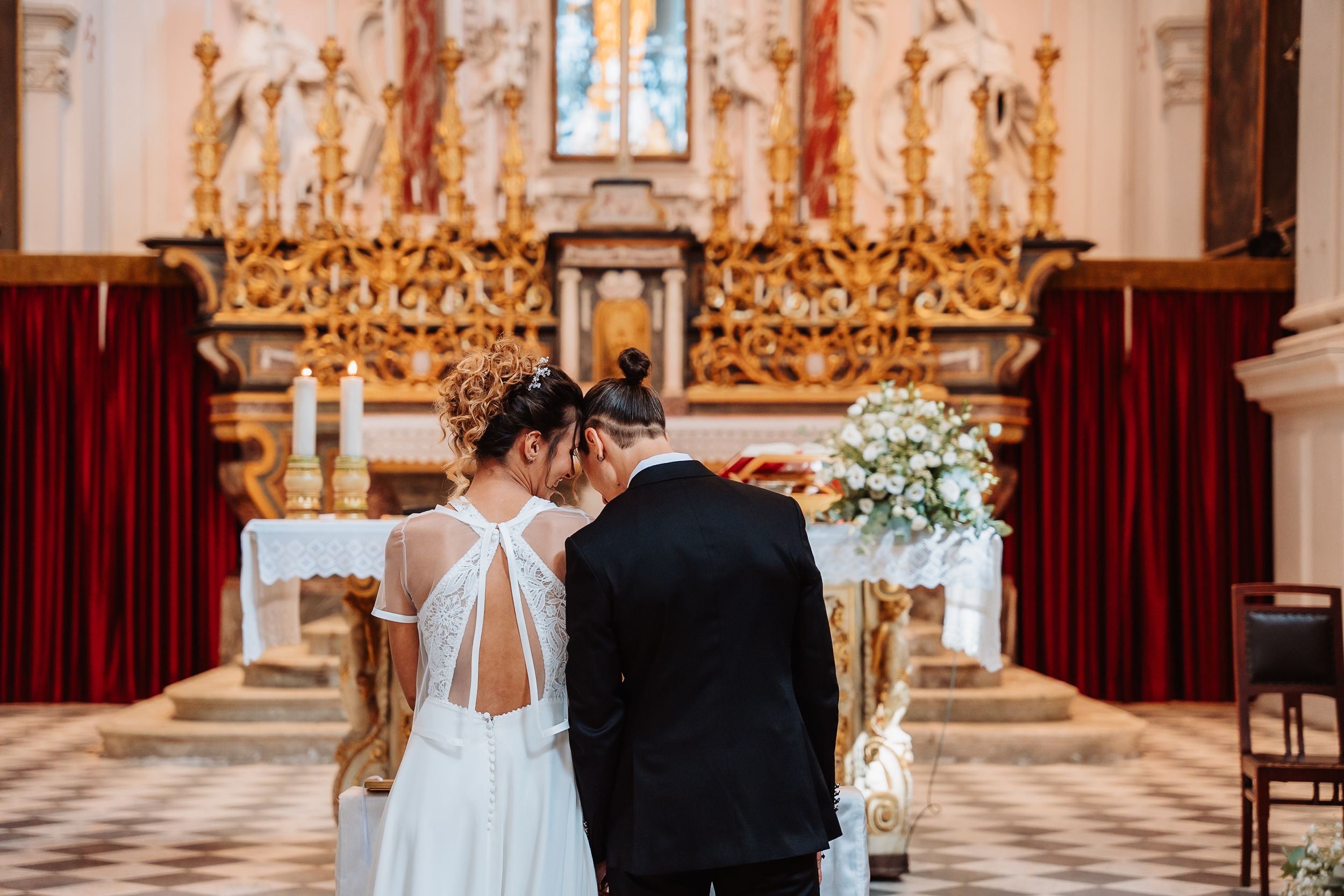 A couple stands at the altar of an ornate church with gold decor and red velvet curtains during their wedding ceremony.