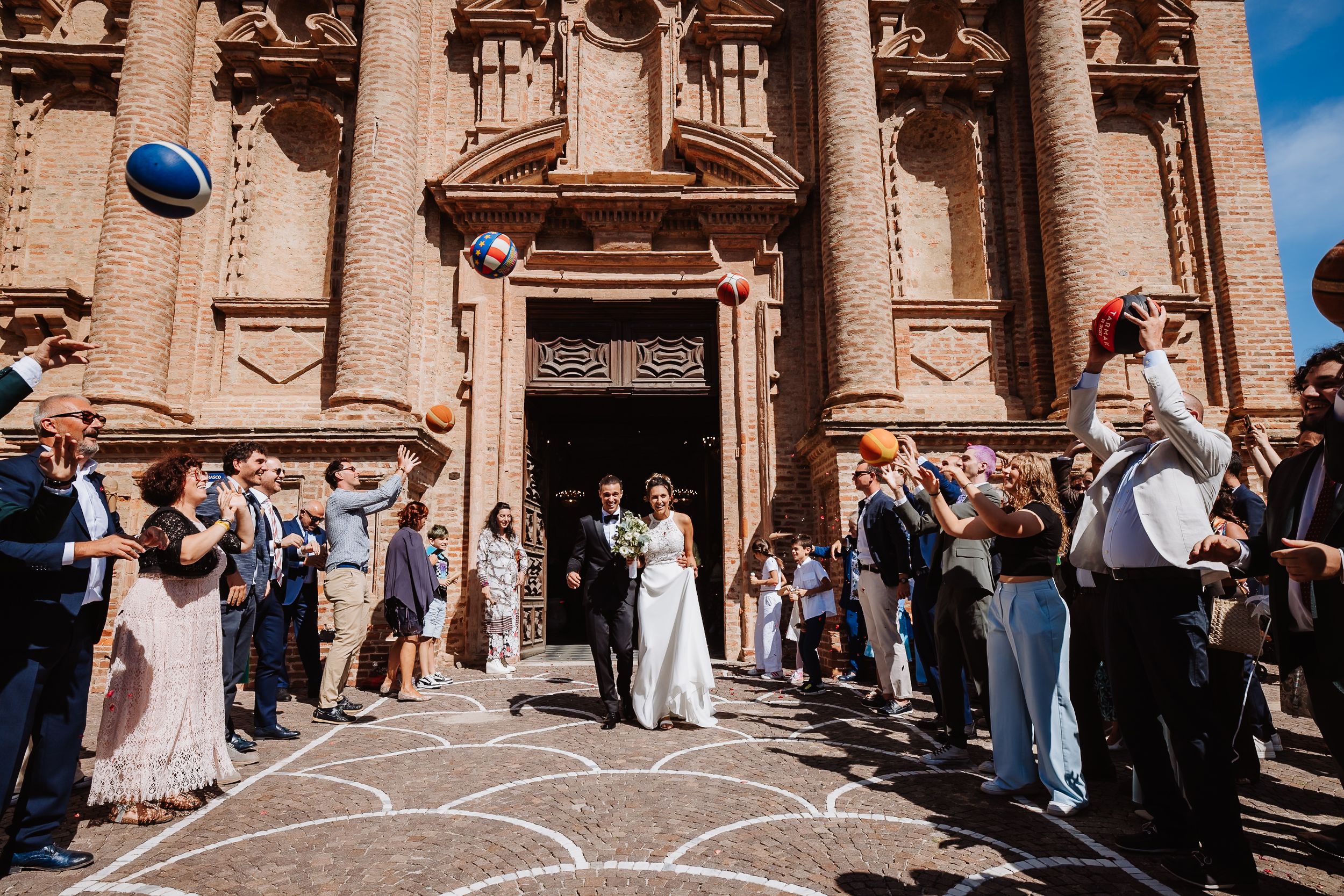 Wedding party gathers outside a grand sandstone church with ornate baroque architecture on a sunny day.