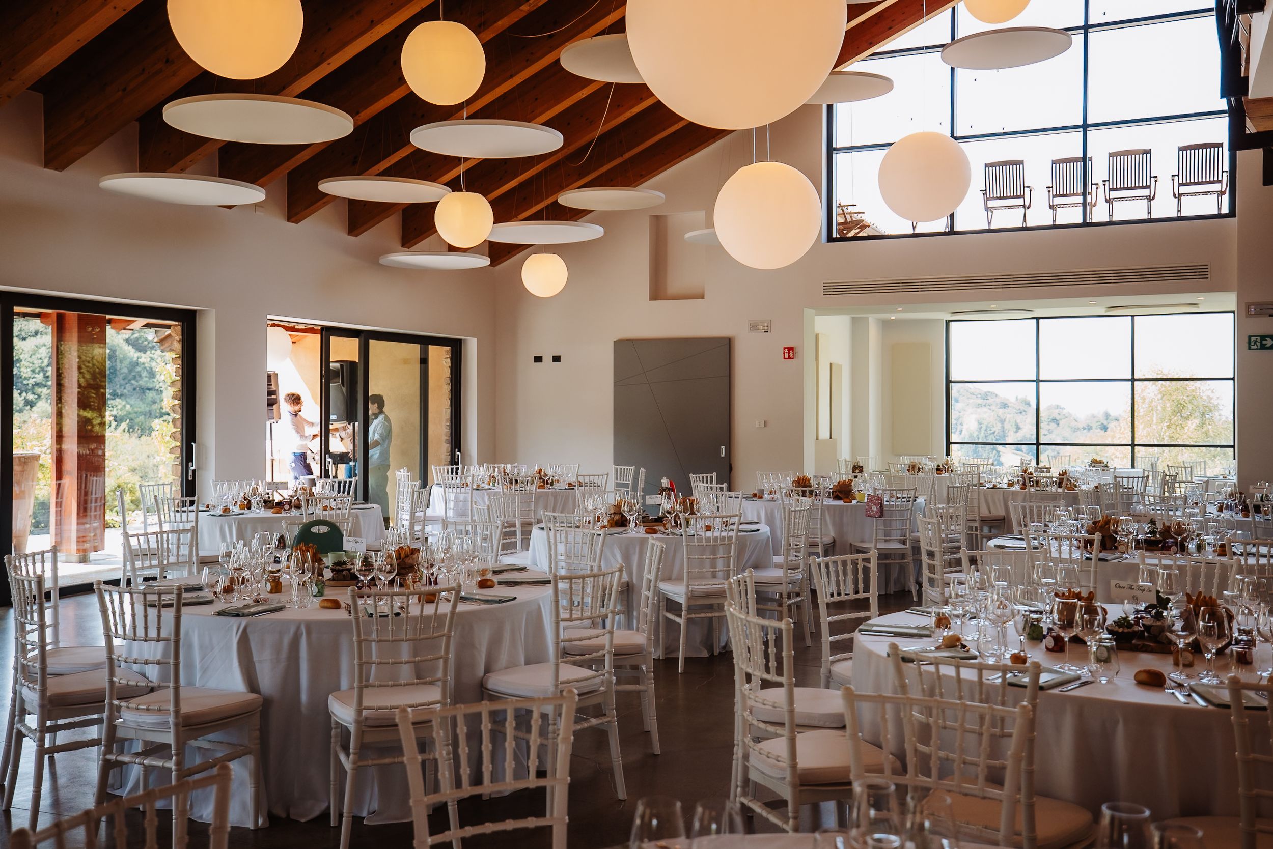 Elegant wedding reception venue with white tables, chiavari chairs, and hanging paper lanterns under wooden beams.