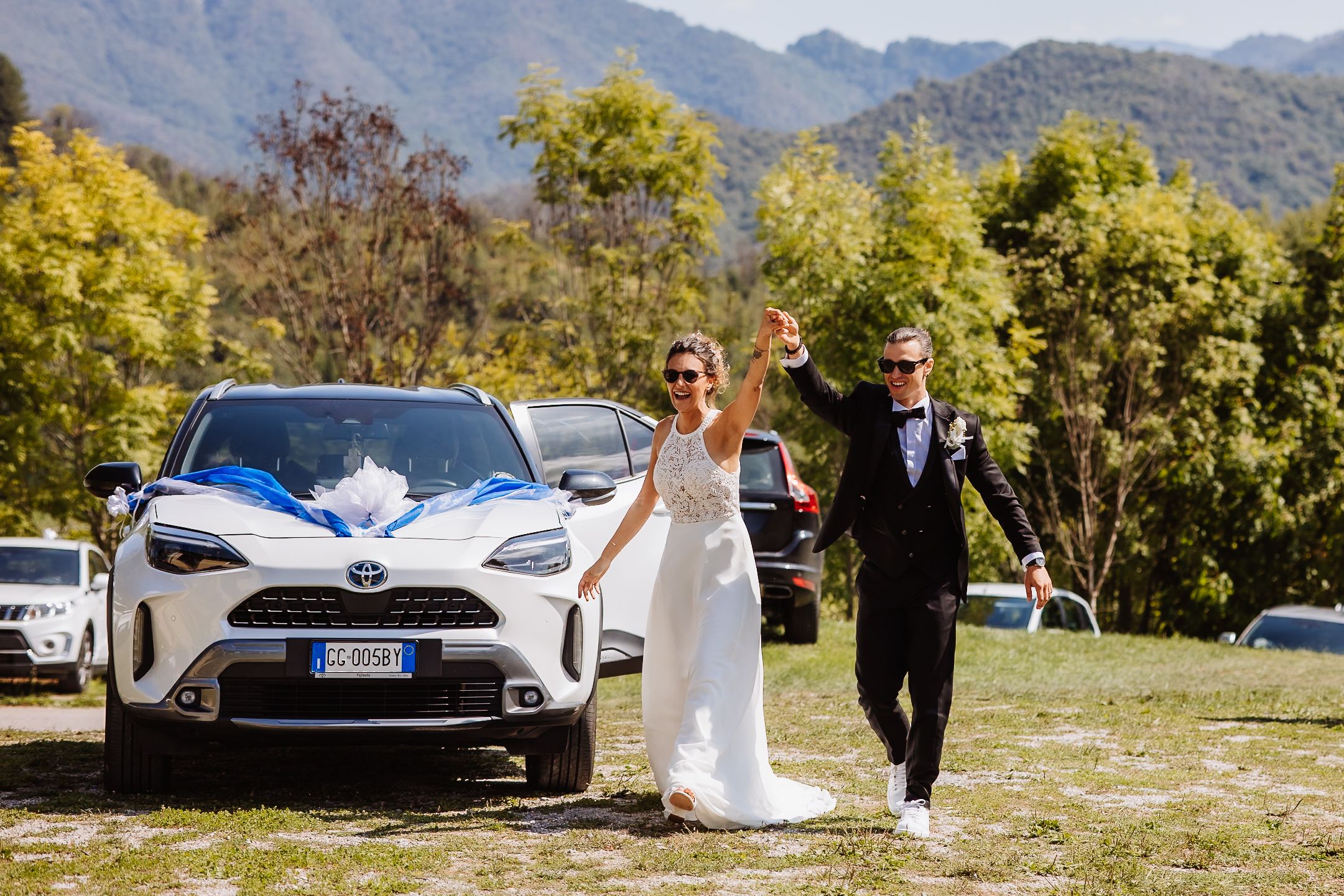 A couple walks together in a scenic mountain landscape with luxury cars parked nearby during a wedding celebration.