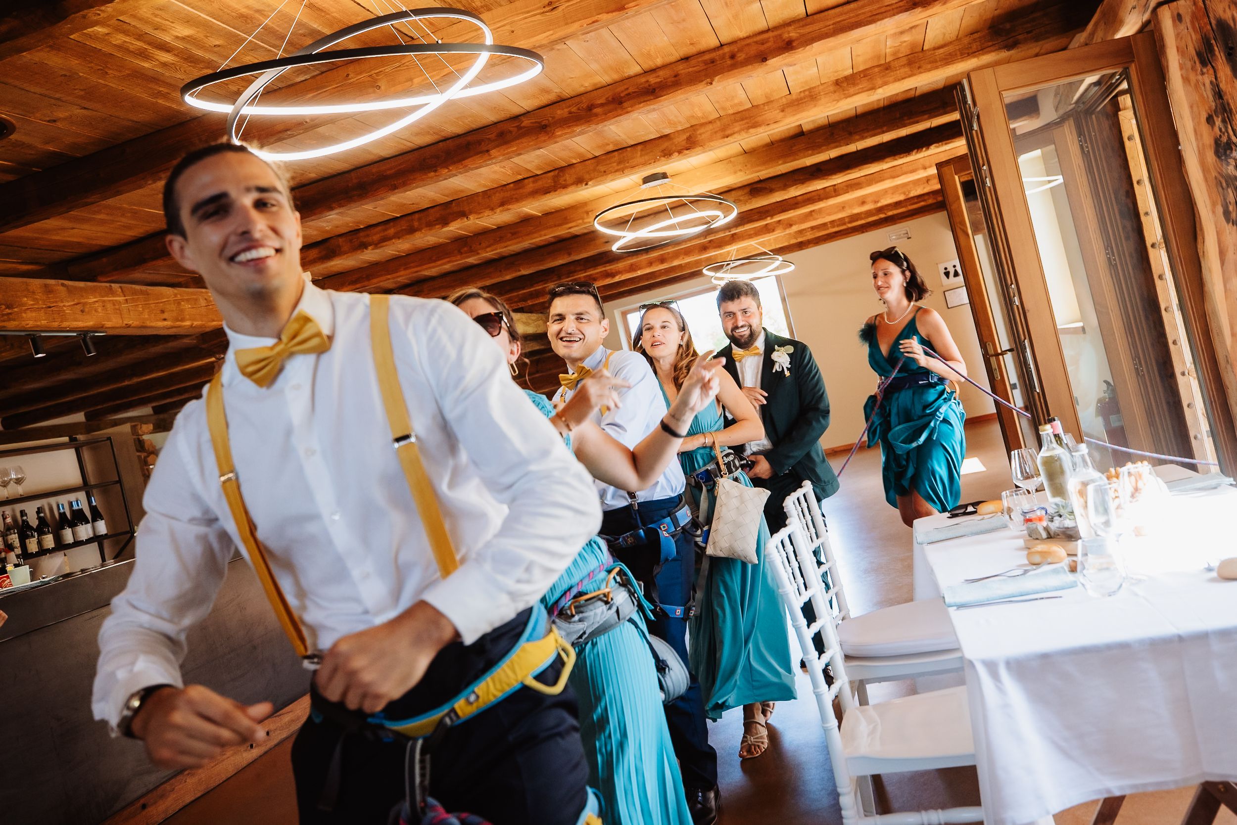 Wedding guests in teal and white attire mingle and dance in a rustic wooden venue with exposed beams.