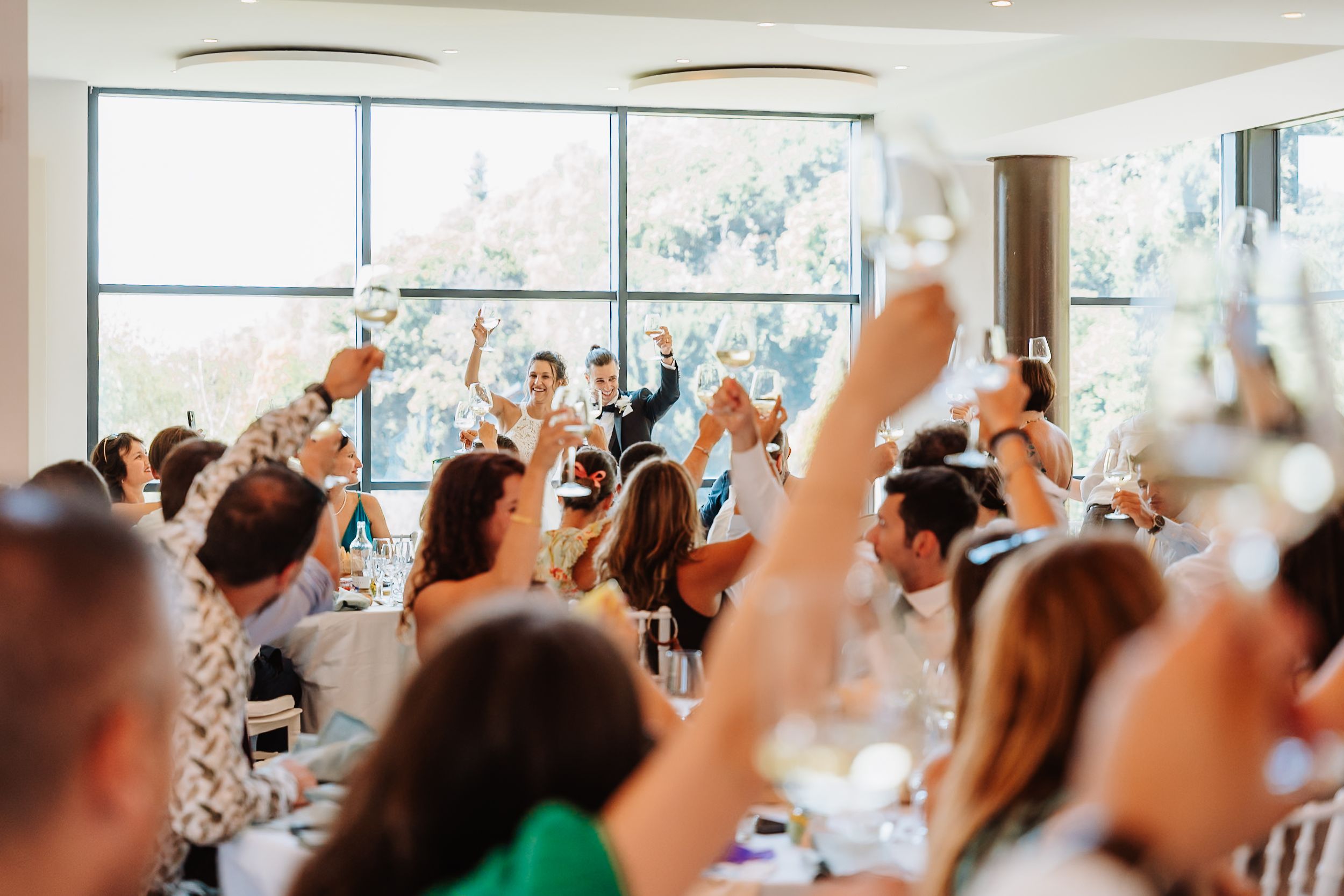 Crowd of wedding guests raise their hands in celebration during a reception with natural light streaming through windows.