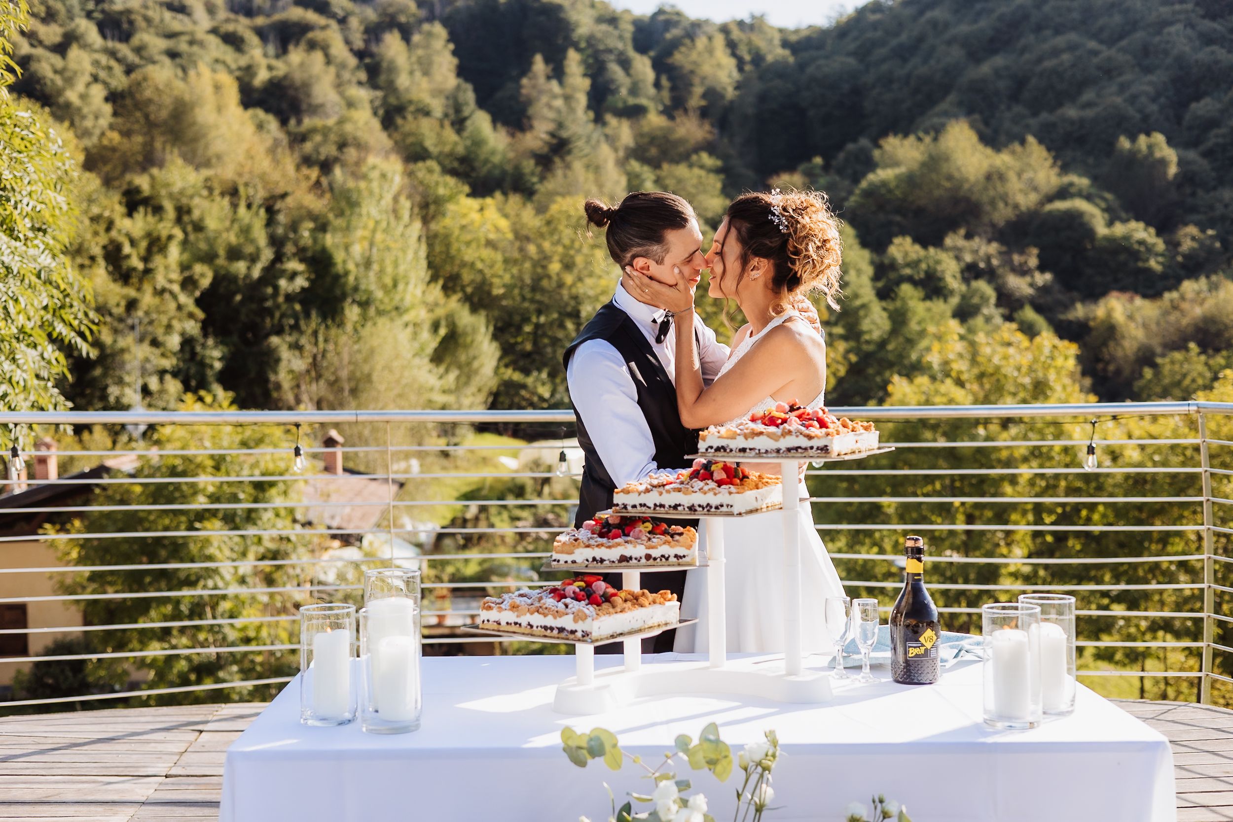 A couple cuts their wedding cake together on an outdoor terrace surrounded by mountains and greenery.