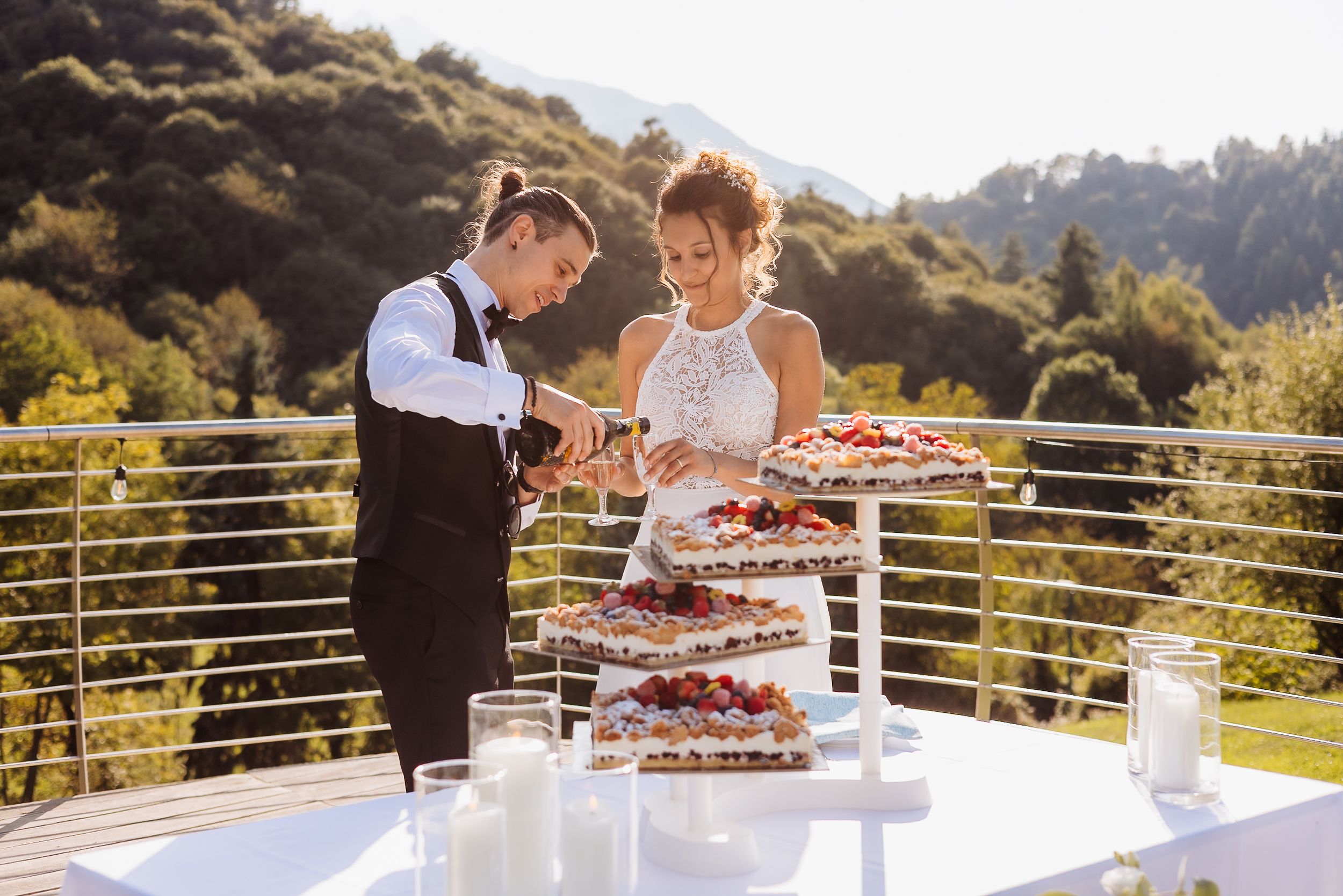 A scenic outdoor wedding reception features guests gathering around a dessert table on a mountain terrace at sunset.