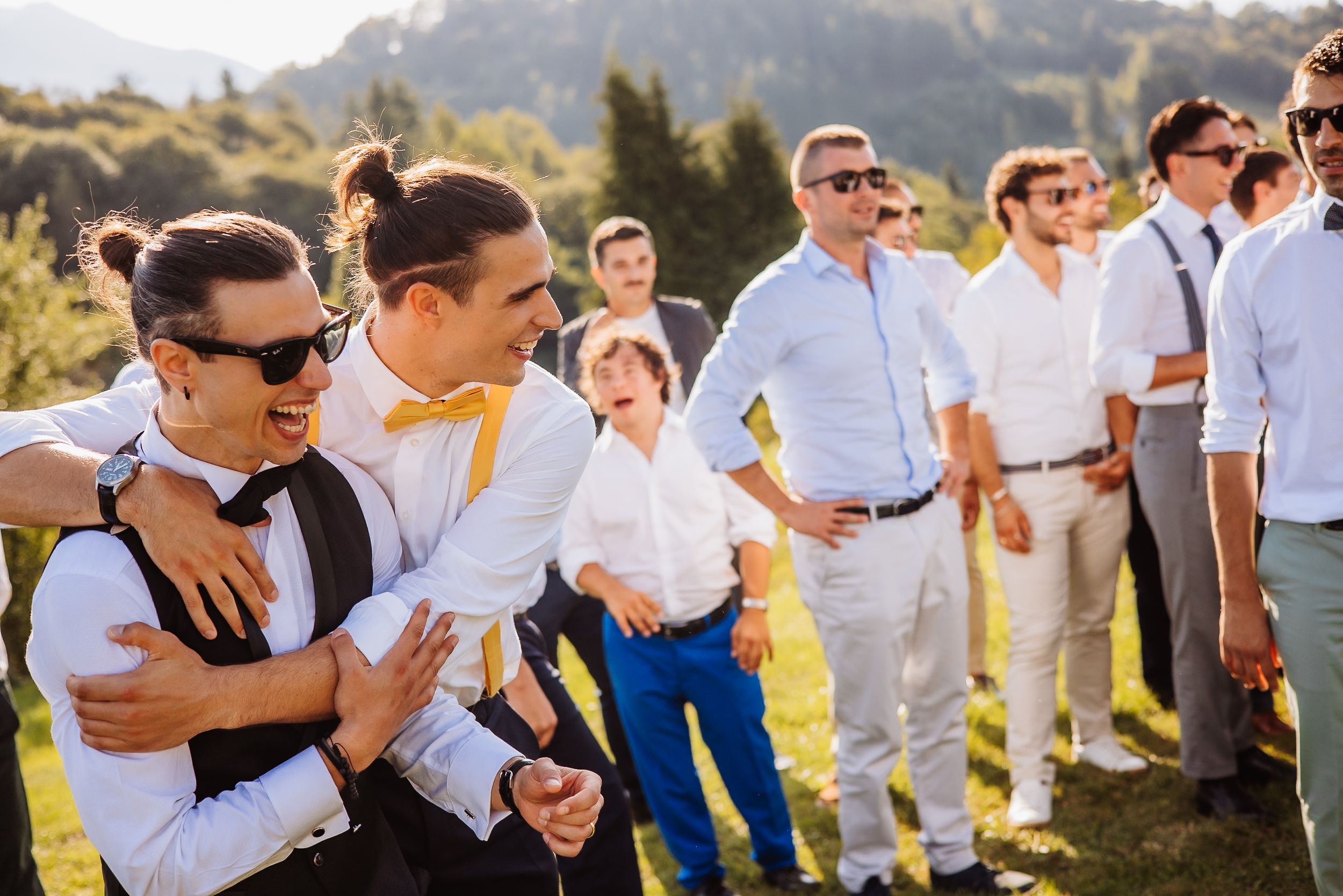 A wedding party in white attire and bow ties celebrates outdoors against a mountainous backdrop on a sunny day.