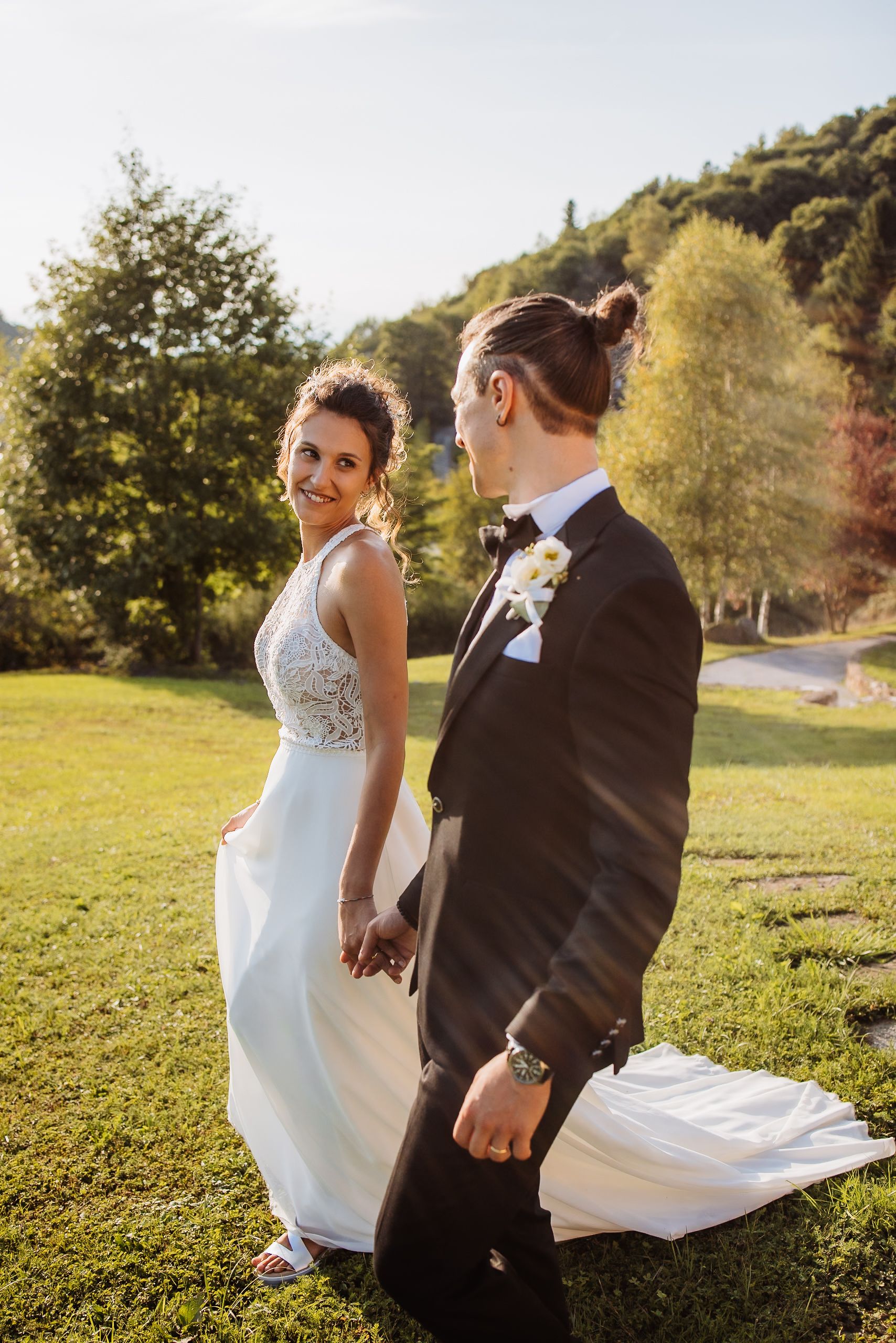 Newlywed couple holding hands in elegant attire stand on sunny grassy hillside during golden hour wedding photoshoot.