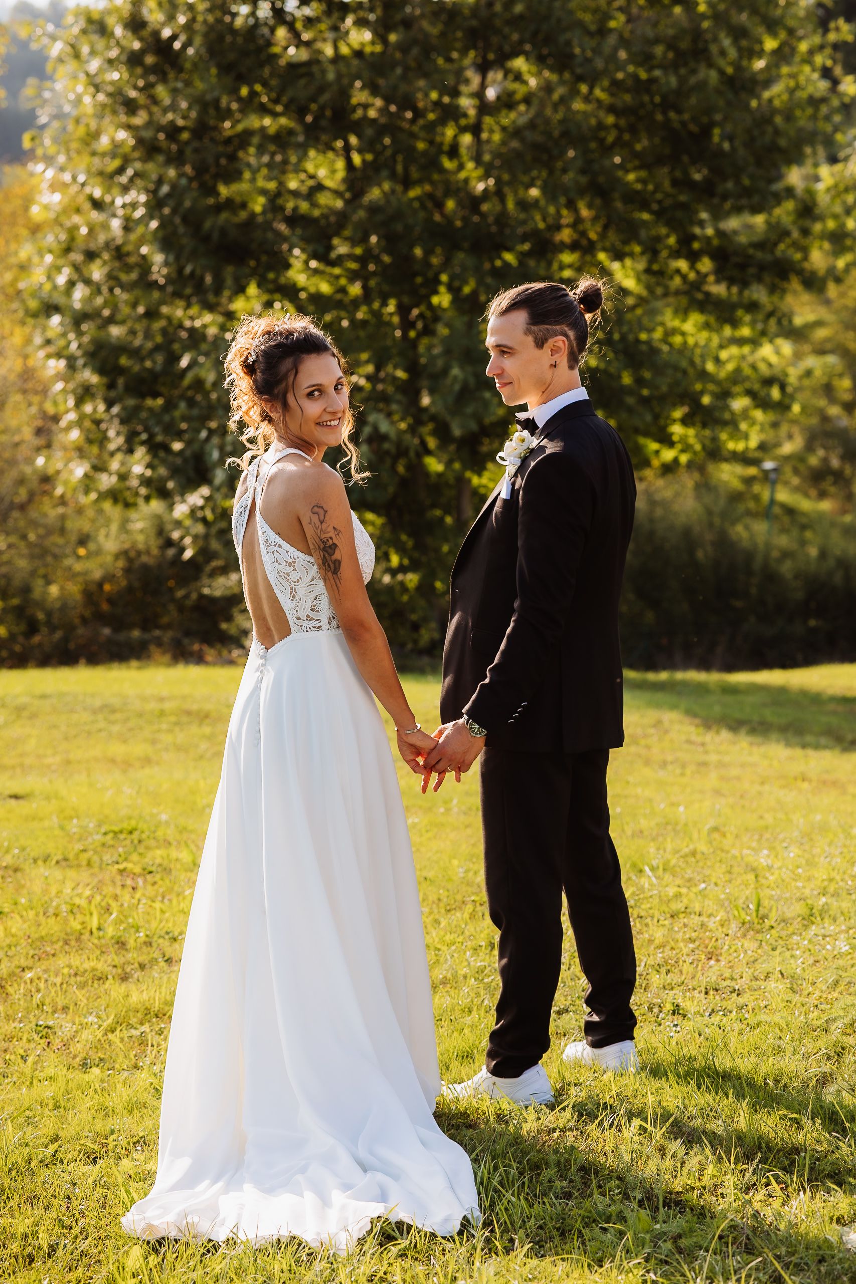 A couple in wedding attire stands together in a sunlit meadow with trees in the background during golden hour.