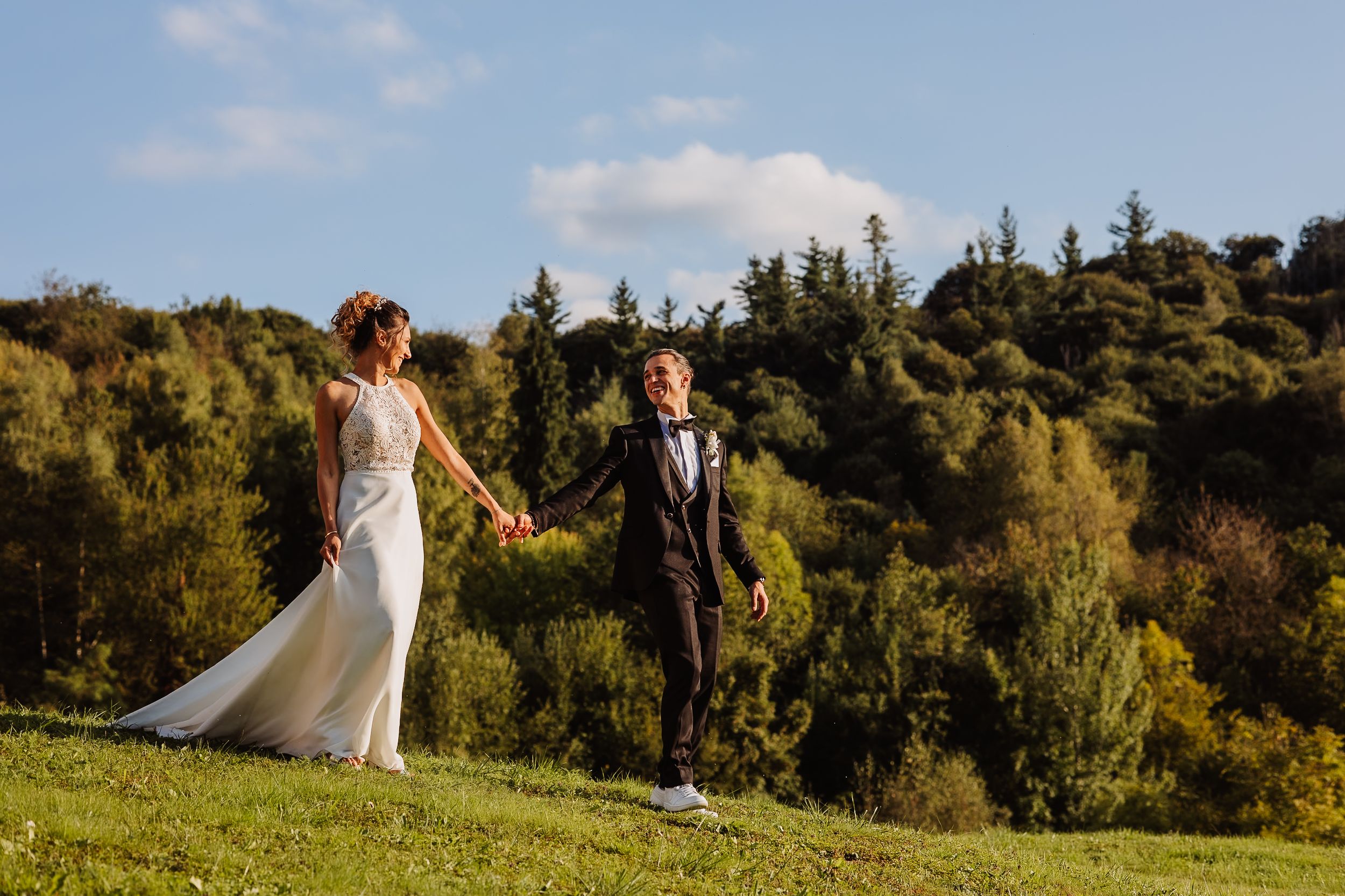 A couple in wedding attire holding hands while standing on a grassy hillside with trees in the background.