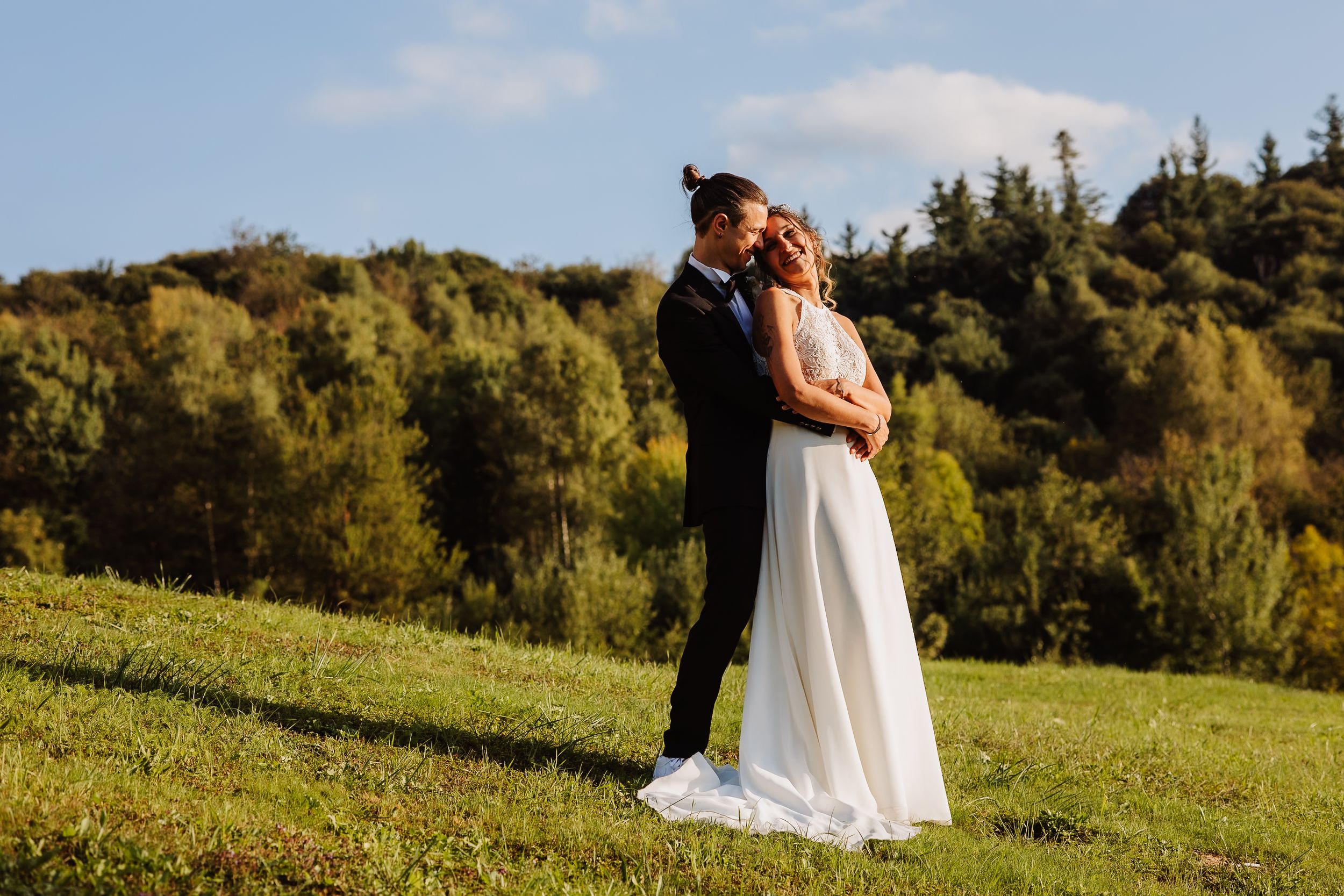 A couple embraces on a grassy hilltop with lush green forest in the background on a sunny afternoon.