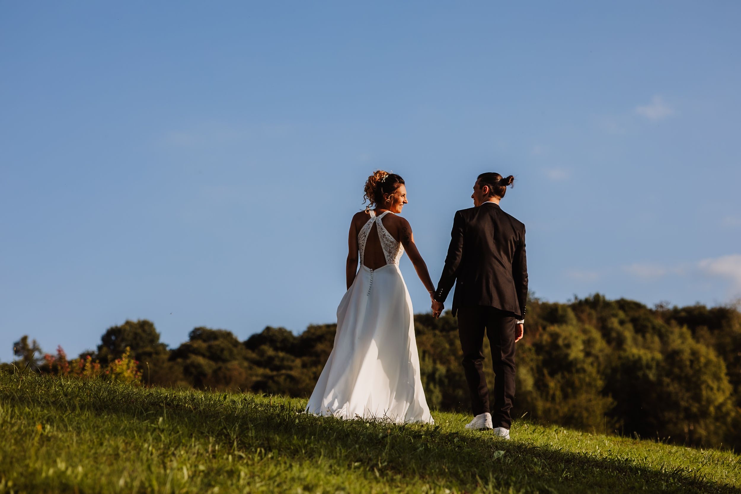 A couple in wedding attire holds hands while standing on a grassy hilltop against a blue sky and trees.