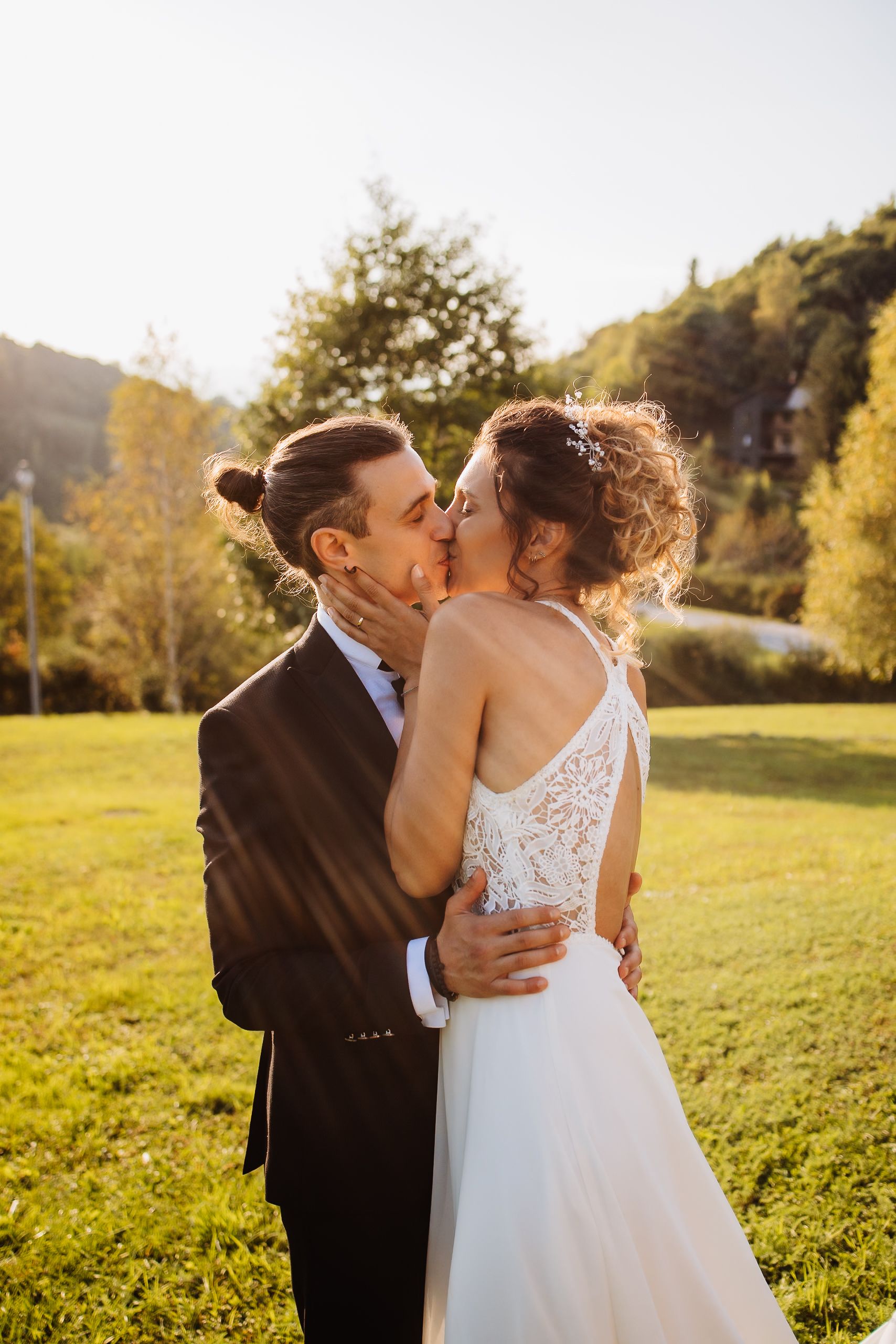A couple shares a romantic kiss in a sunlit meadow with autumn trees and mountains in the background.