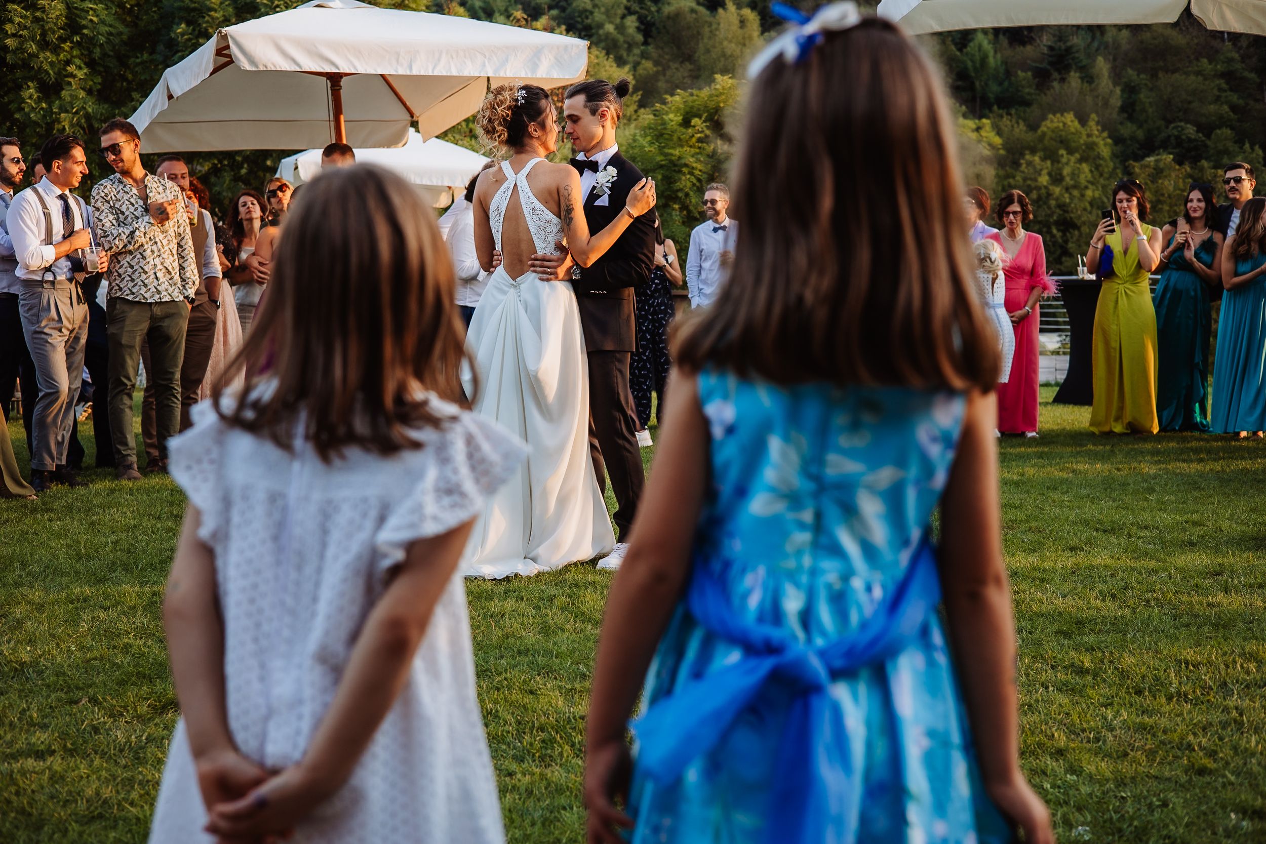 Wedding guests mingle and socialize on a sunny lawn with white umbrellas during an outdoor reception.