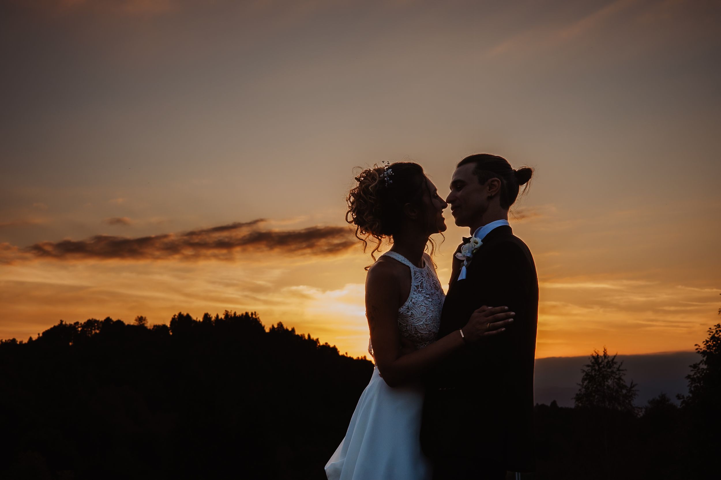Silhouettes of a couple embrace against a dramatic orange and purple sunset sky with trees in the background.
