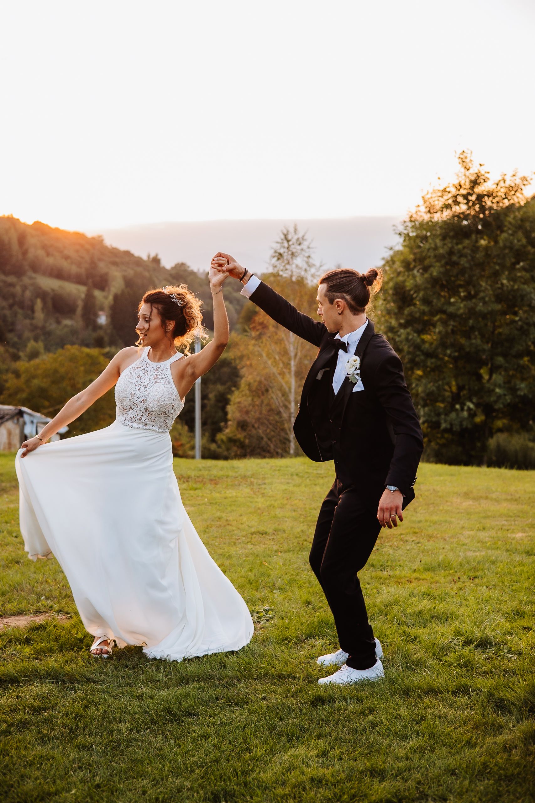 A newlywed couple dances joyfully on a grassy hilltop at sunset during their wedding celebration.