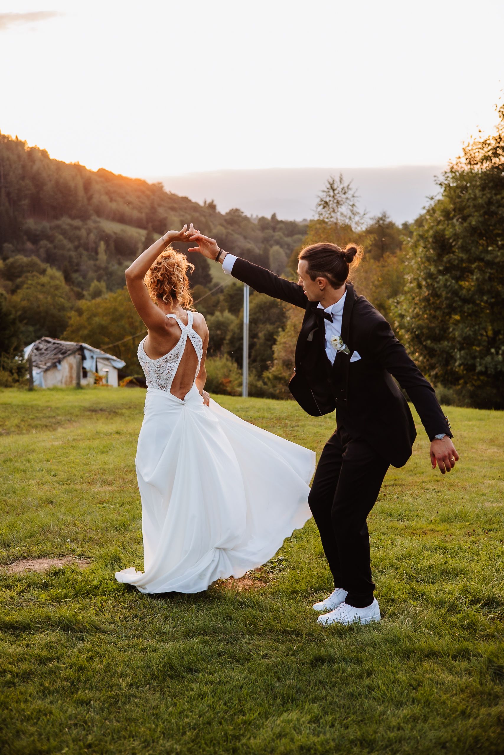 A couple in wedding attire performs martial arts kicks together on a grassy hillside at sunset.