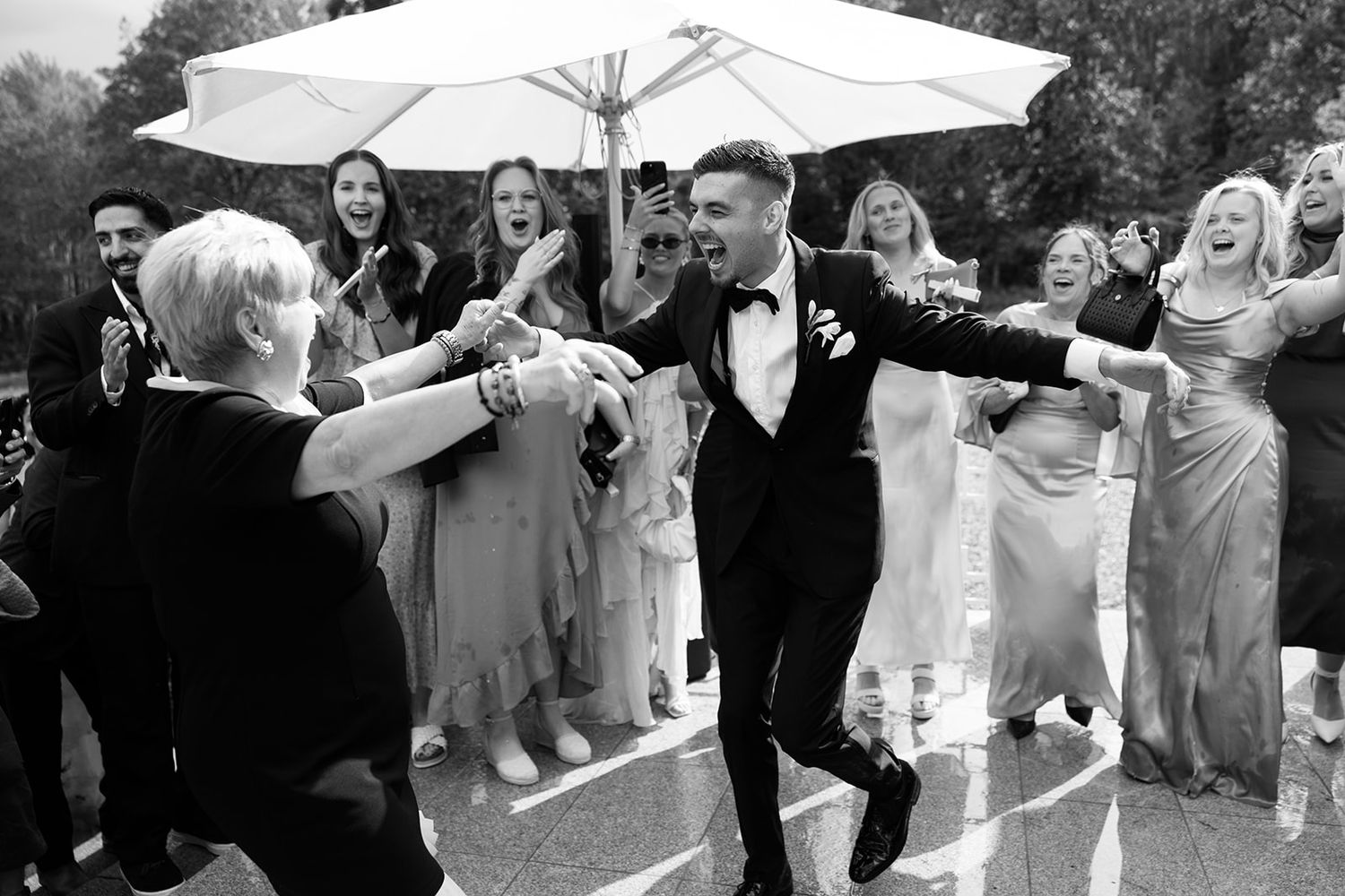 Groomsmen in black tuxedos pose for a fun photo while kneeling and laughing together on a stone patio.