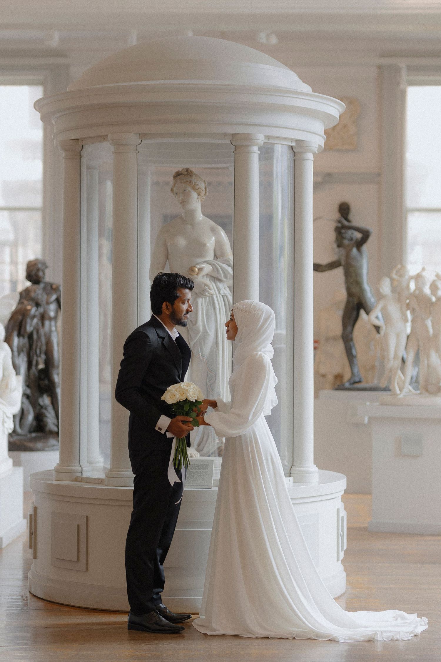 A couple stands together in an elegant white room filled with classical sculptures and architectural columns.