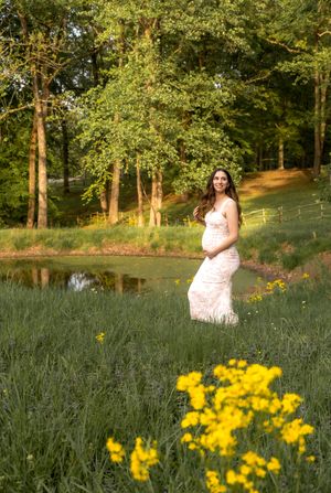 Expecting mother in white lace dress poses among lavender and yellow flowers in a sunlit meadow.