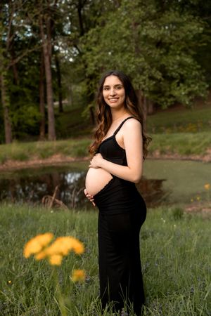 A maternity photoshoot in a natural outdoor setting with yellow wildflowers and a pond in the background.