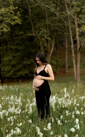 An expectant mother stands in a field of white wildflowers surrounded by lush green trees.