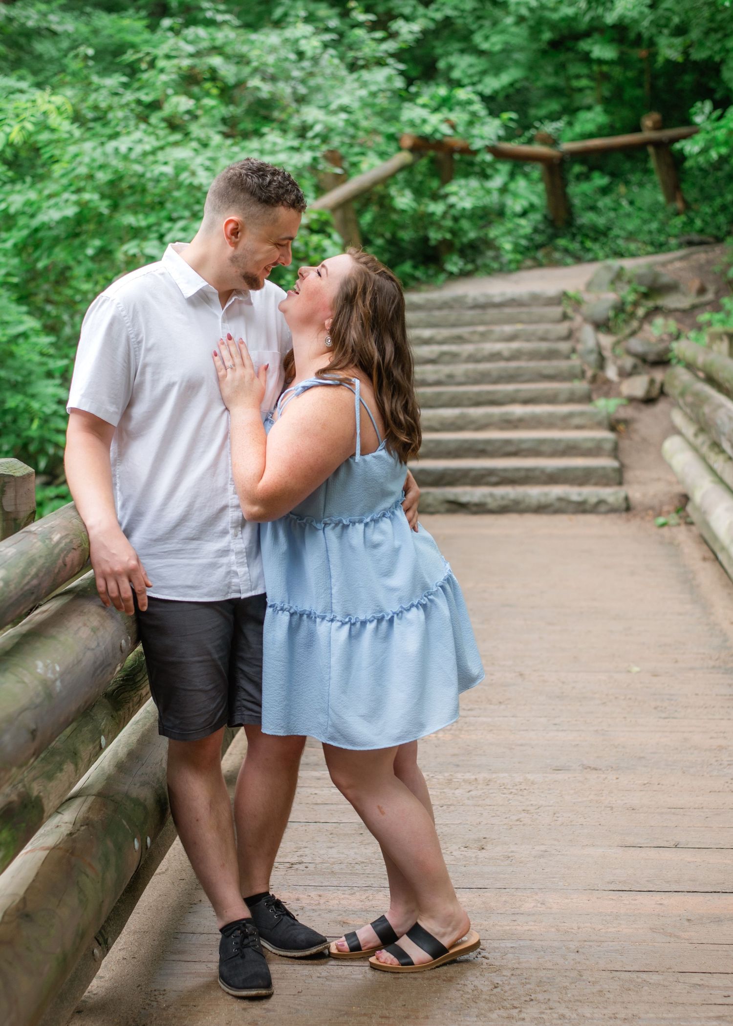 A couple shares romantic moments on a wooden bridge pathway surrounded by lush green foliage.