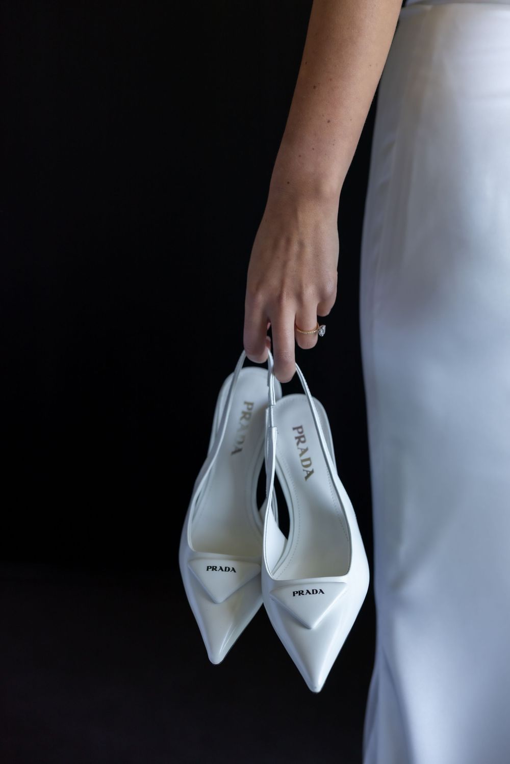 Hand holding elegant white Prada pointed-toe shoes against dark background.